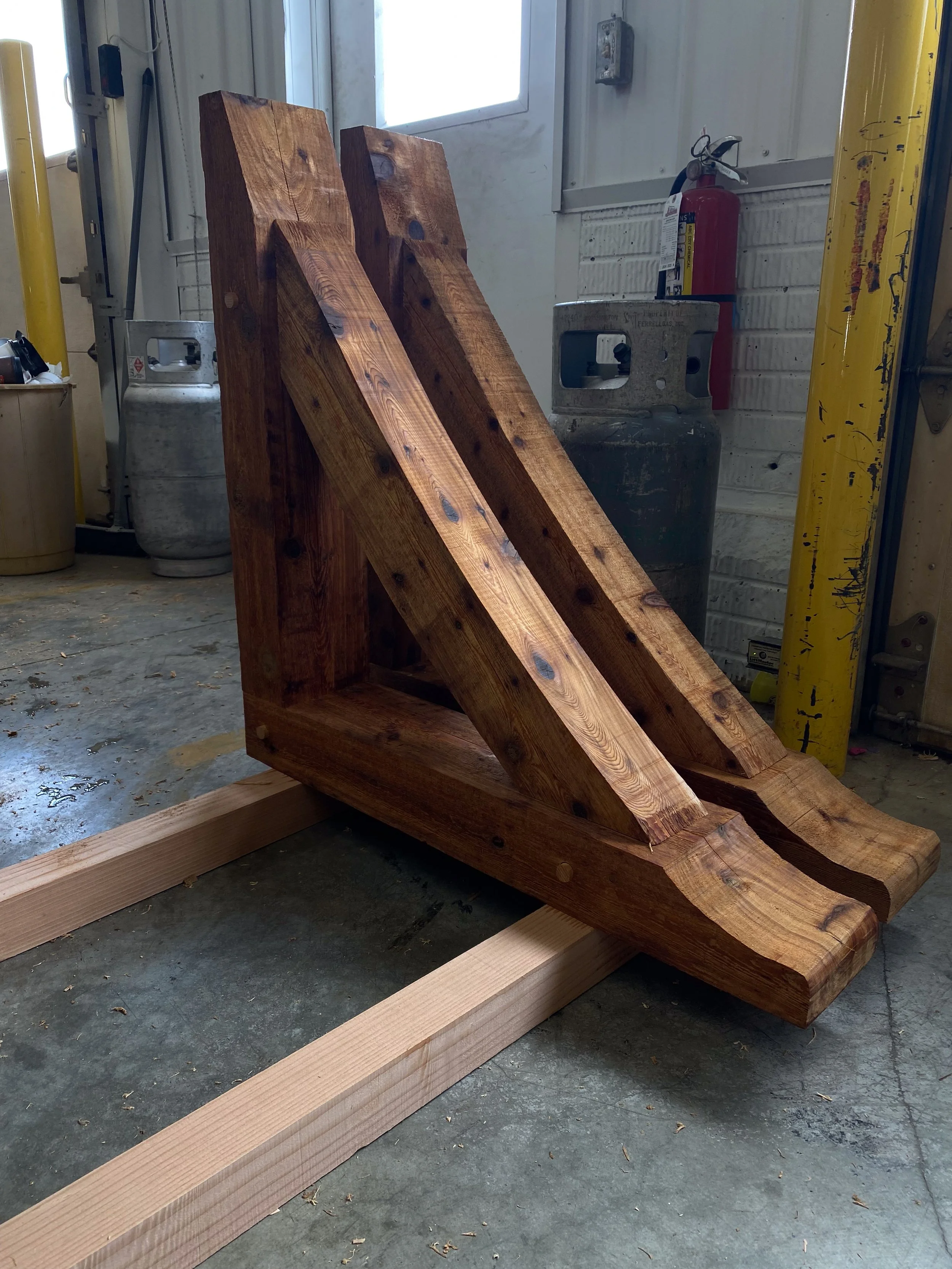 A large, unfinished wooden sawhorse in a workshop with concrete floor, a yellow post, a fire extinguisher, and propane tanks in the background.