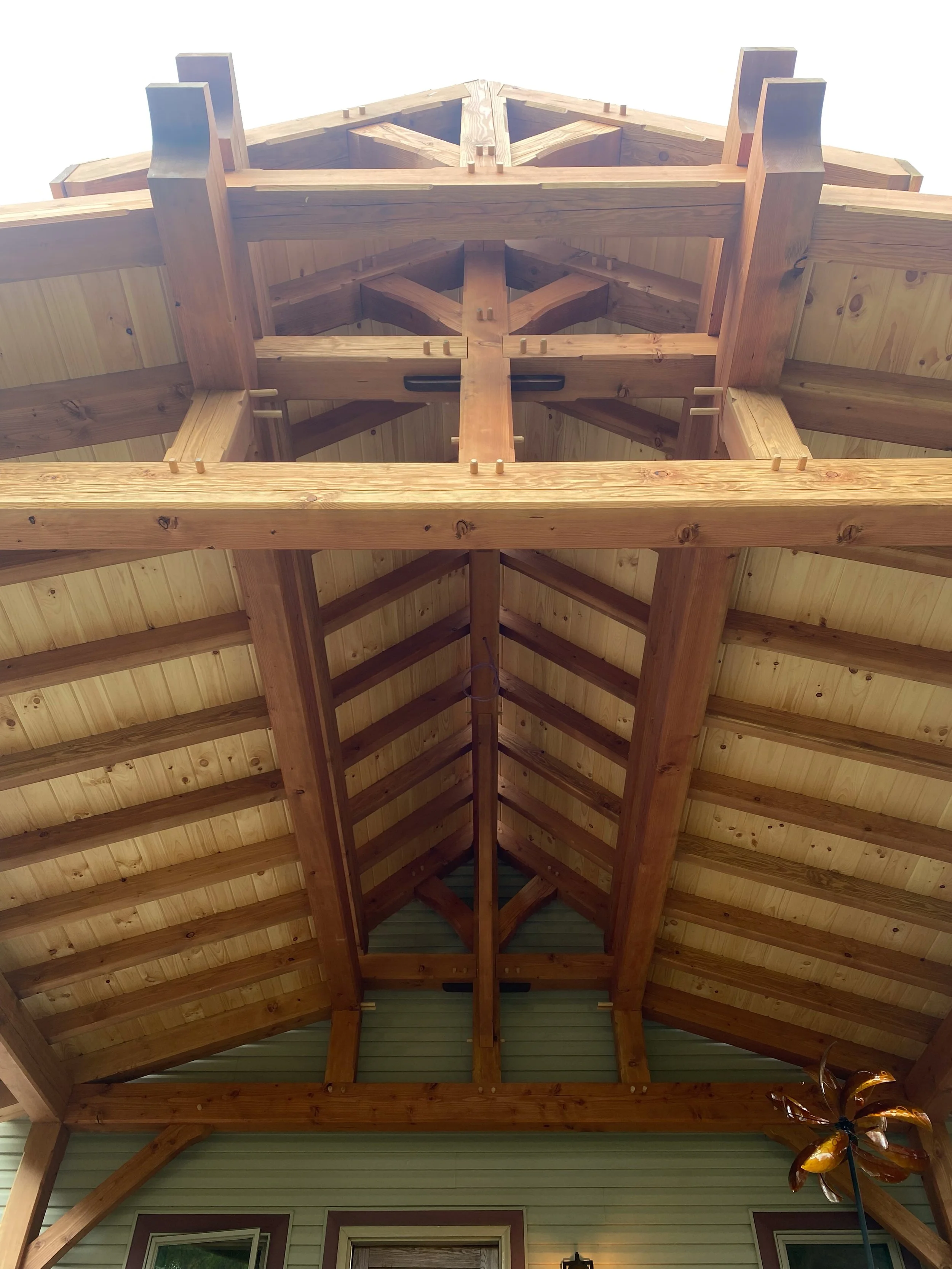 Looking up at a wooden roof structure with exposed beams inside a building or porch, with part of a house's wall and a decorative windmill visible at the bottom right.