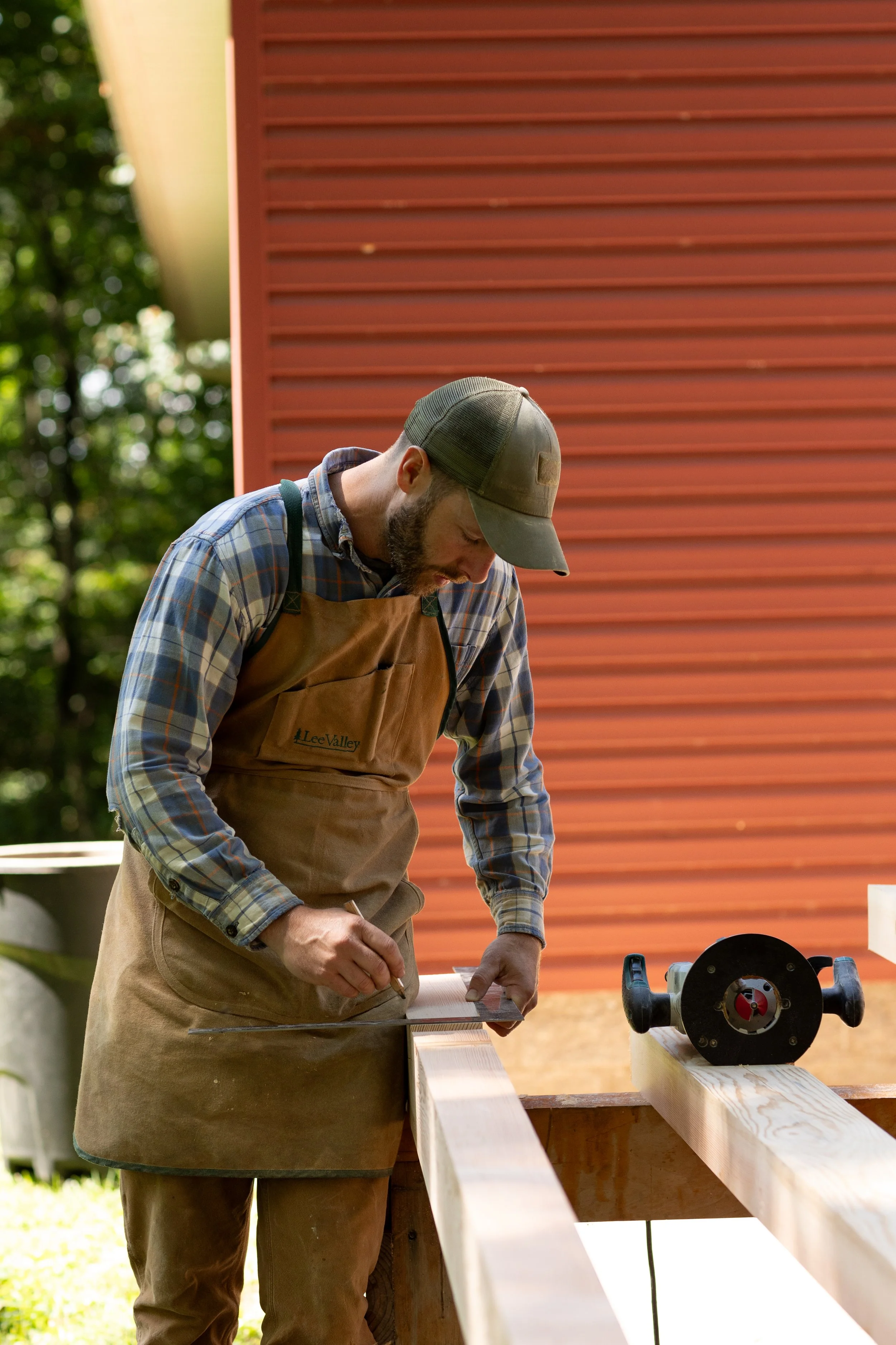 Man wearing a plaid shirt and a brown apron with a green cap working on a woodworking project outdoors with a bow saw on the wooden beam.