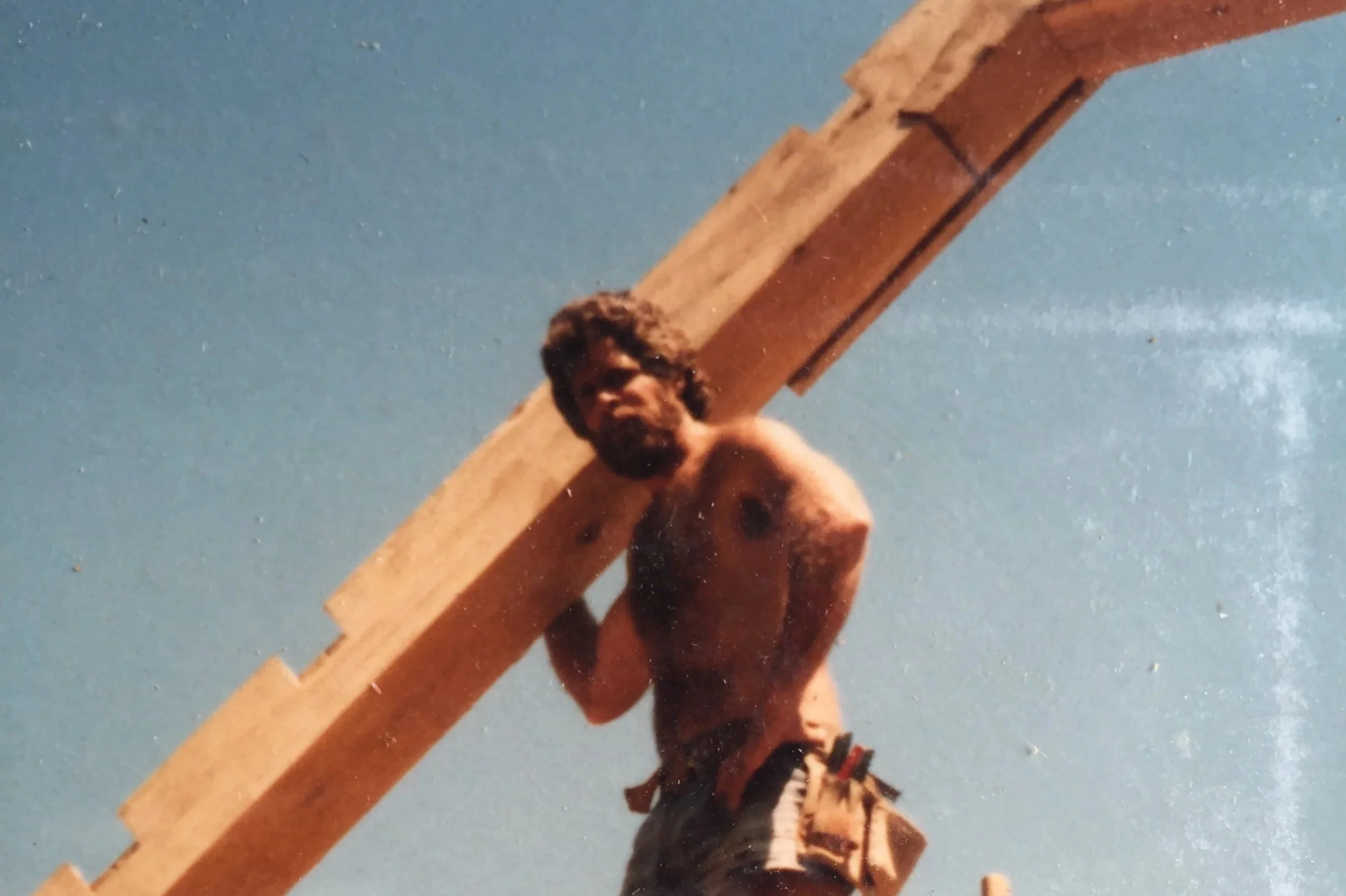 Man with curly hair and beard, shirtless, carrying a large wooden cross on his shoulder against a blue sky.