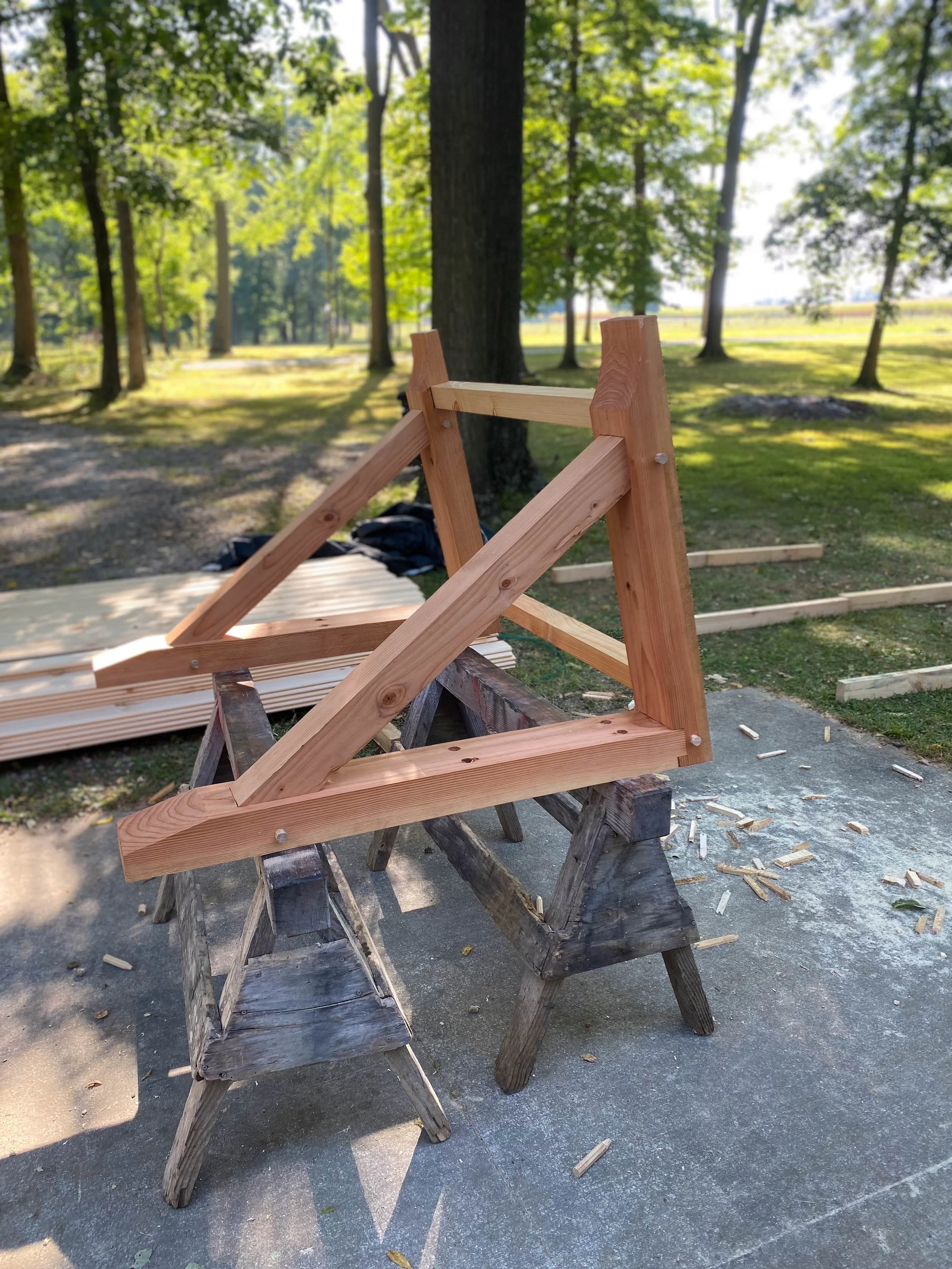 A wooden sawhorse on a workbench outdoors, with more wood planks and sawdust nearby, set in a forested area with green trees and sunlight filtering through.