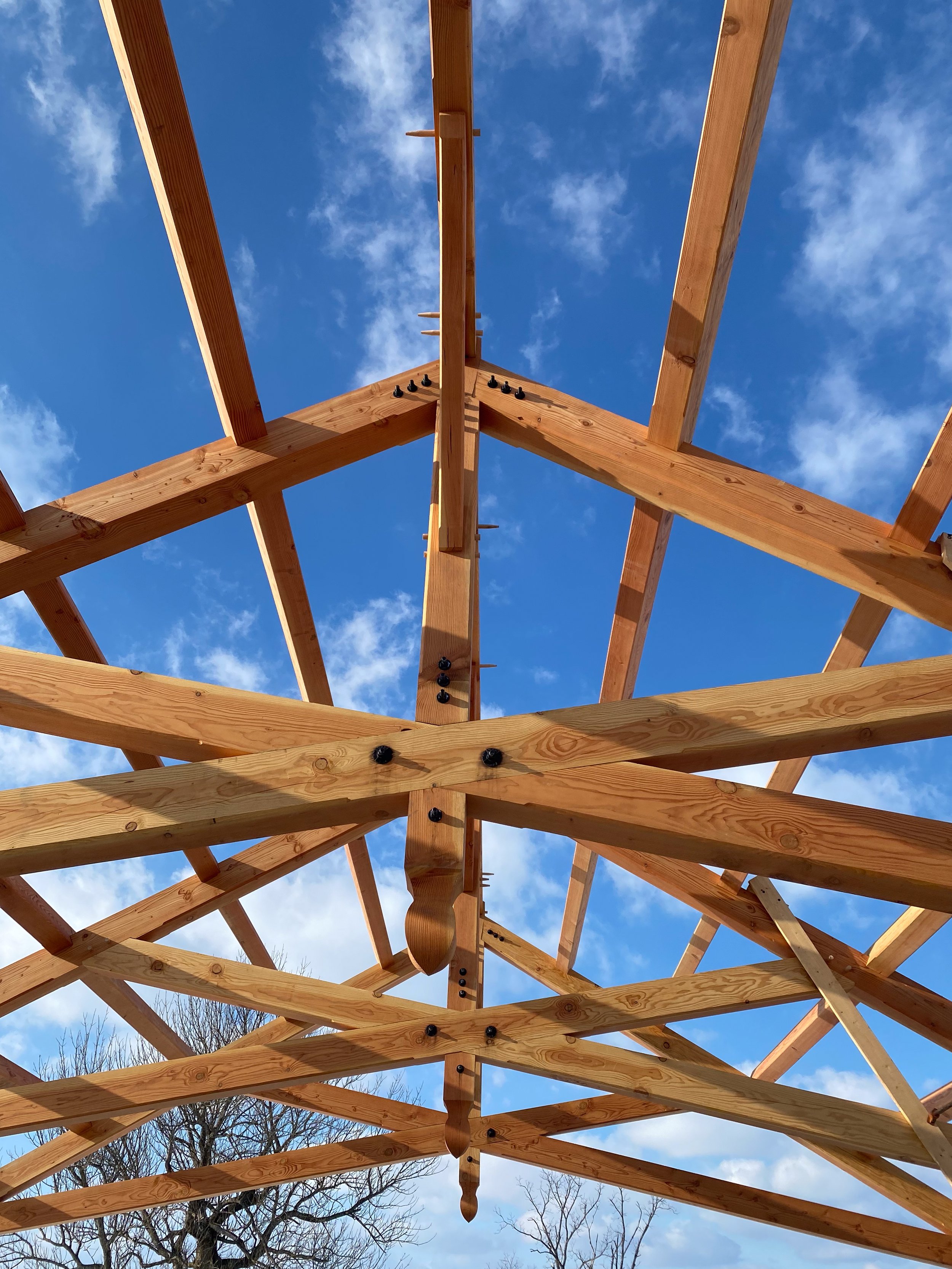 View of a wooden roof framing structure under a blue sky with scattered clouds, showing the beams and joints.