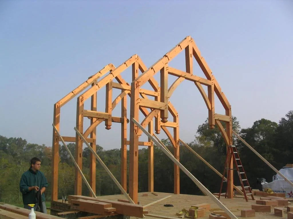 Wooden framing for a house under construction, with a ladder and construction materials on site, and a man standing nearby.