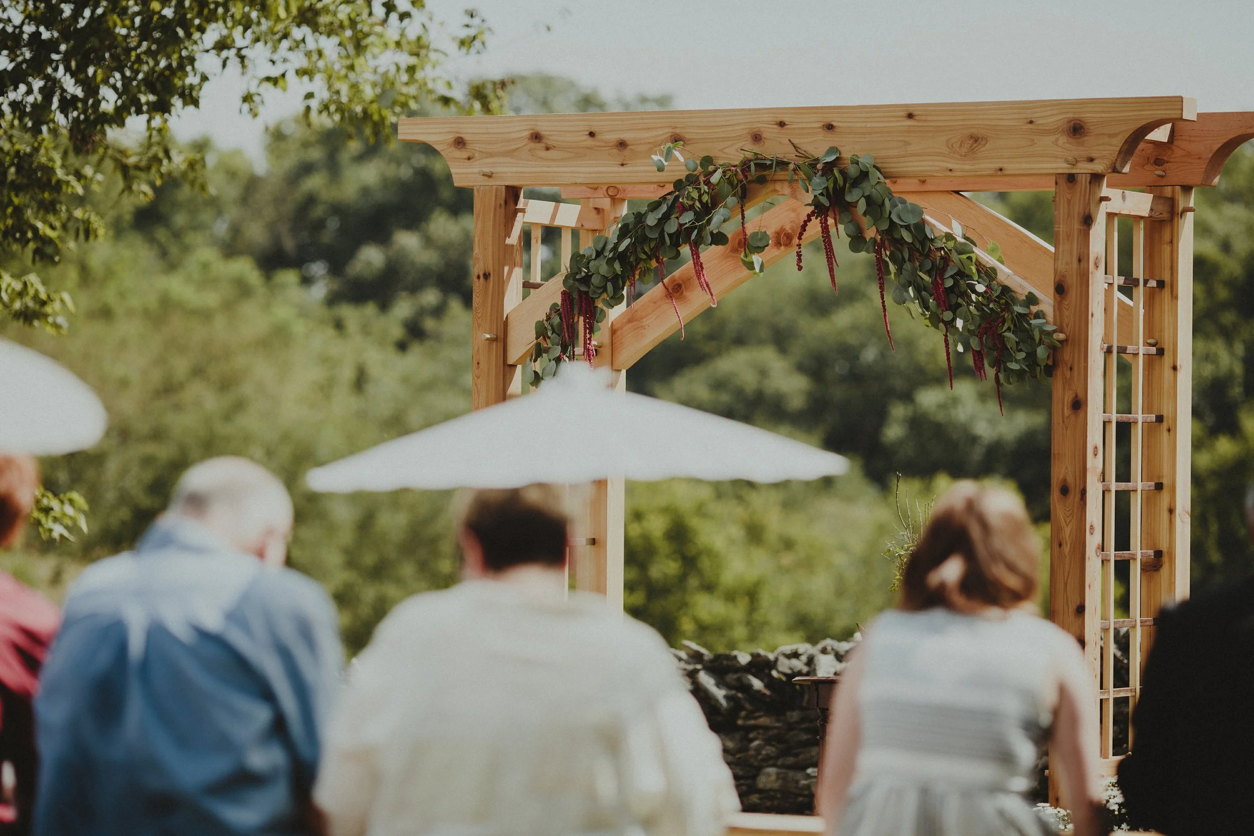 Outdoor wedding ceremony with guests seated facing a wooden arch decorated with greenery and red flowers, and a white umbrella providing shade.