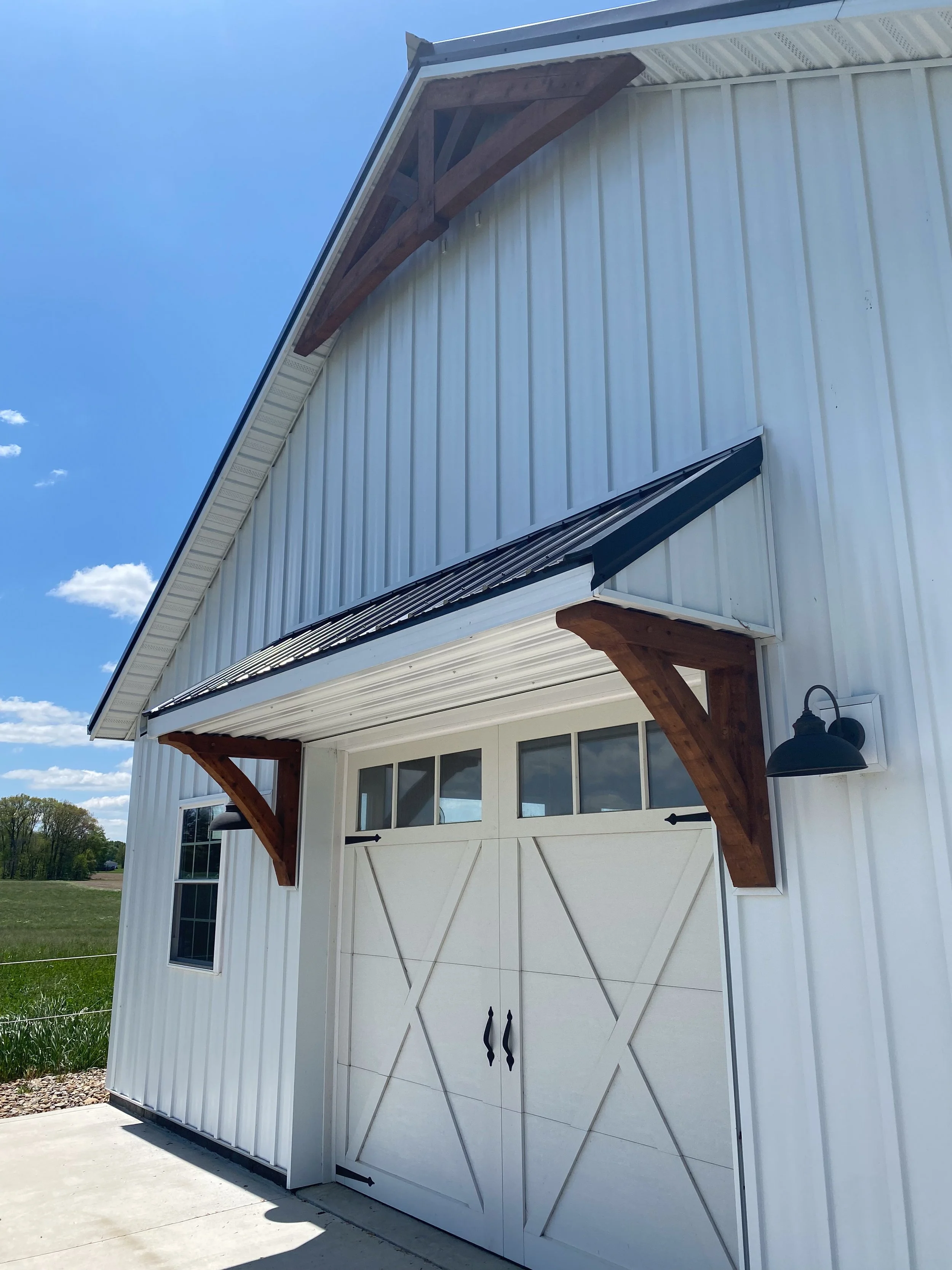White barn-style garage with a metal roof, decorative wooden supports under the eaves, a black light fixture outside, and glass-paneled garage doors, set against a blue sky with a few clouds.