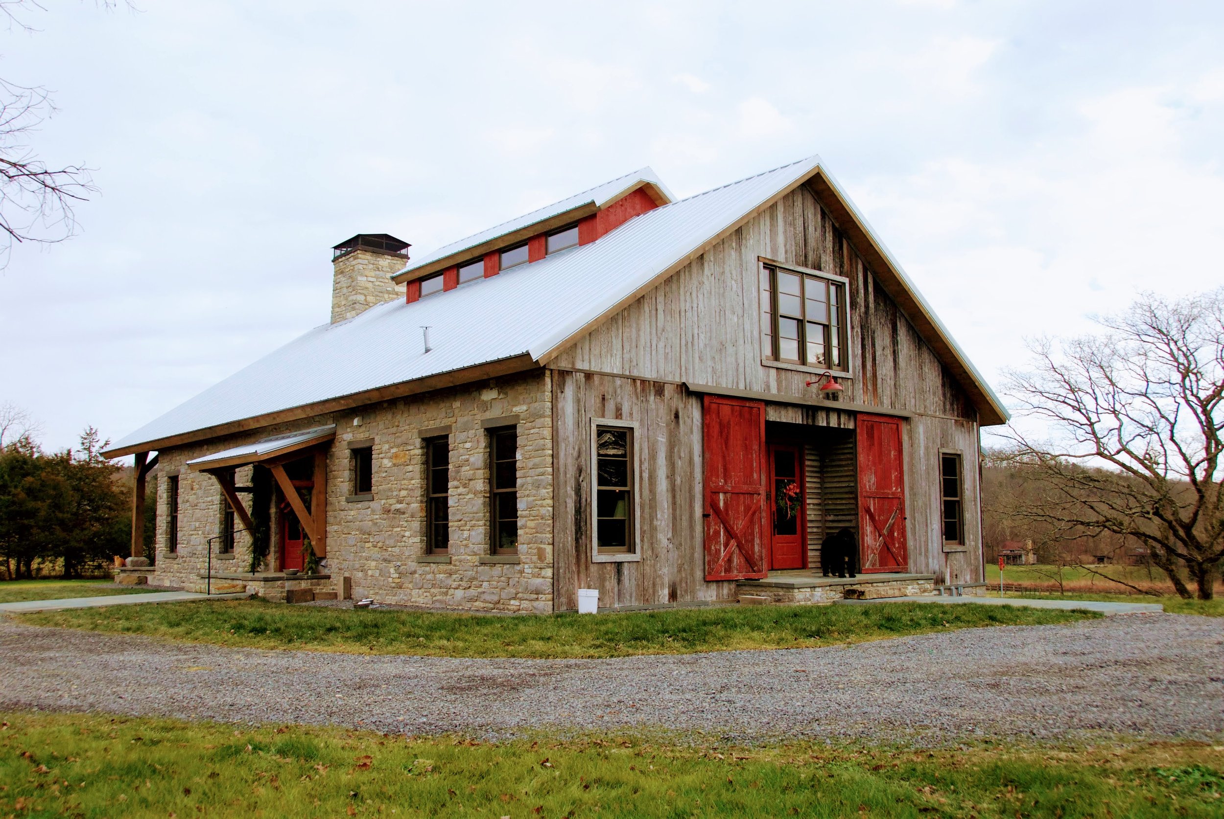 A rustic farmhouse with stone and weathered wood exterior, red barn doors, and a white metal roof, surrounded by trees and grassy areas.