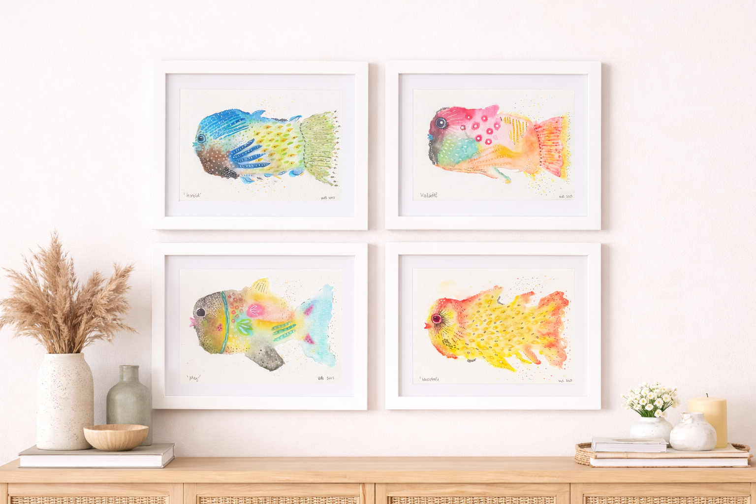 Four framed watercolor fish art prints hanging on a white wall above a wooden sideboard. The artwork features colorful, abstract fish paintings. The sideboard decor includes vases, a small bowl, and books, with a bunch of dried pampas grass in a larg
