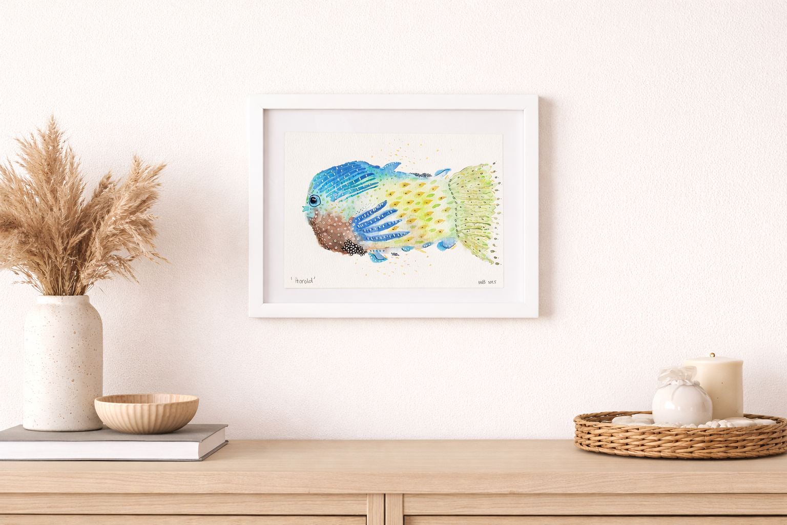 Watercolor illustration of a colorful tropical fish framed and hanging on a white wall above a wooden sideboard, with decorative items including a vase with dried grasses, a bowl, and candles.