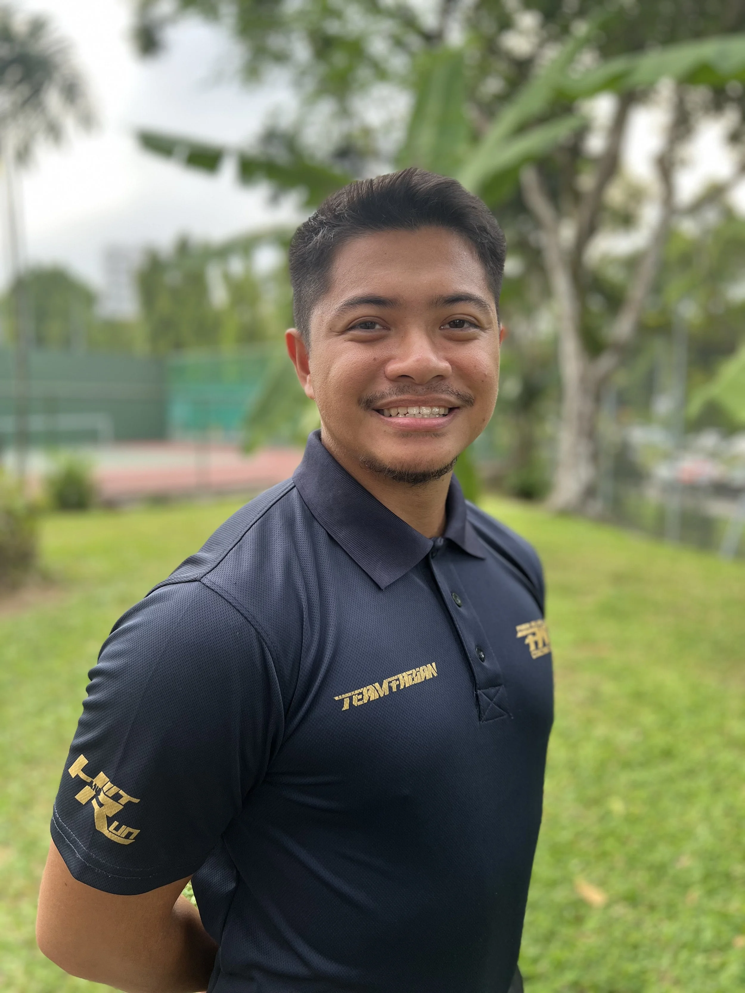 Young woman smiling outdoors, wearing a navy blue polo shirt with logos on the chest and sleeve, with green foliage in the background.