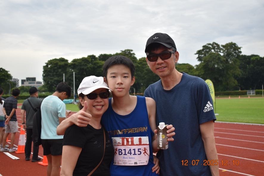 Three people, two adults and one young boy, at a running track during a race event. The woman on the left is smiling and giving a thumbs-up, the boy in the middle is wearing a blue running outfit with a bib number B1415, and the man on the right is smiling and holding a water bottle. The background shows other participants, track cones, and trees under a cloudy sky.