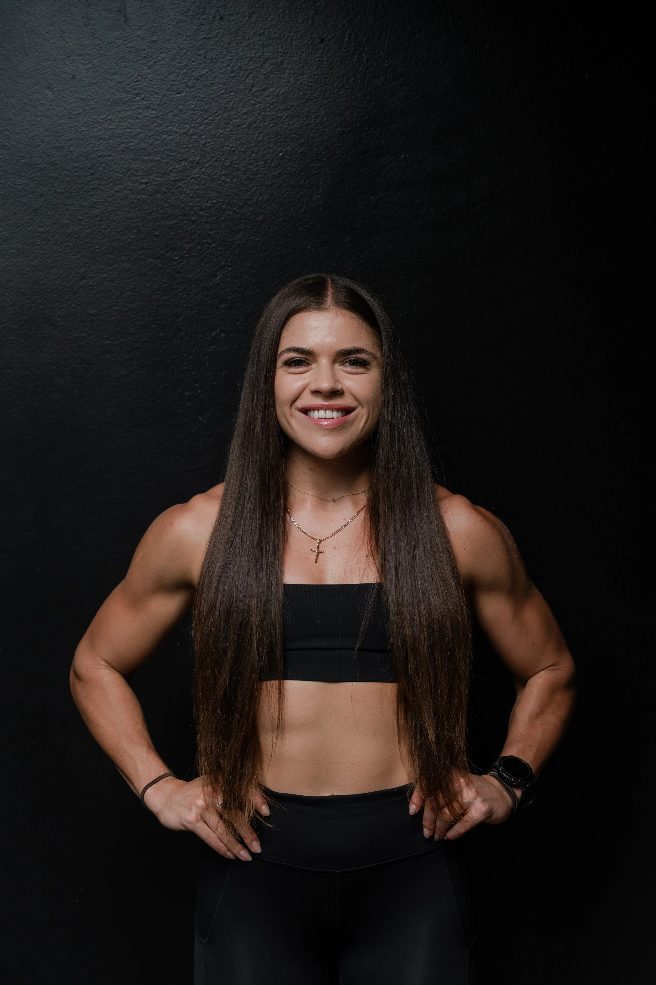 A smiling woman with long brown hair, wearing a black sports bra and black workout leggings, standing with hands on hips against a black textured wall.