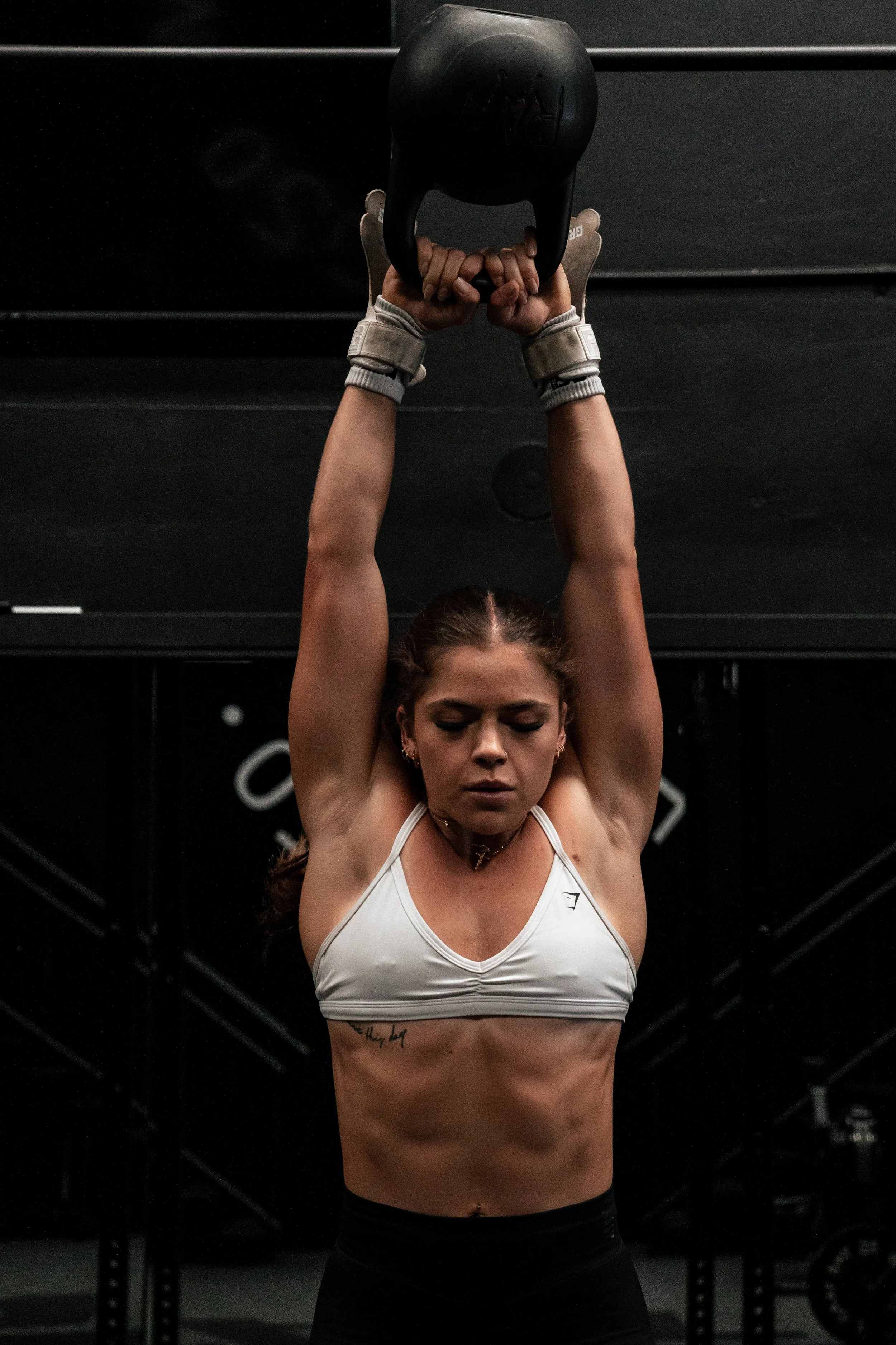 A woman in athletic wear lifting a kettlebell overhead at the gym.