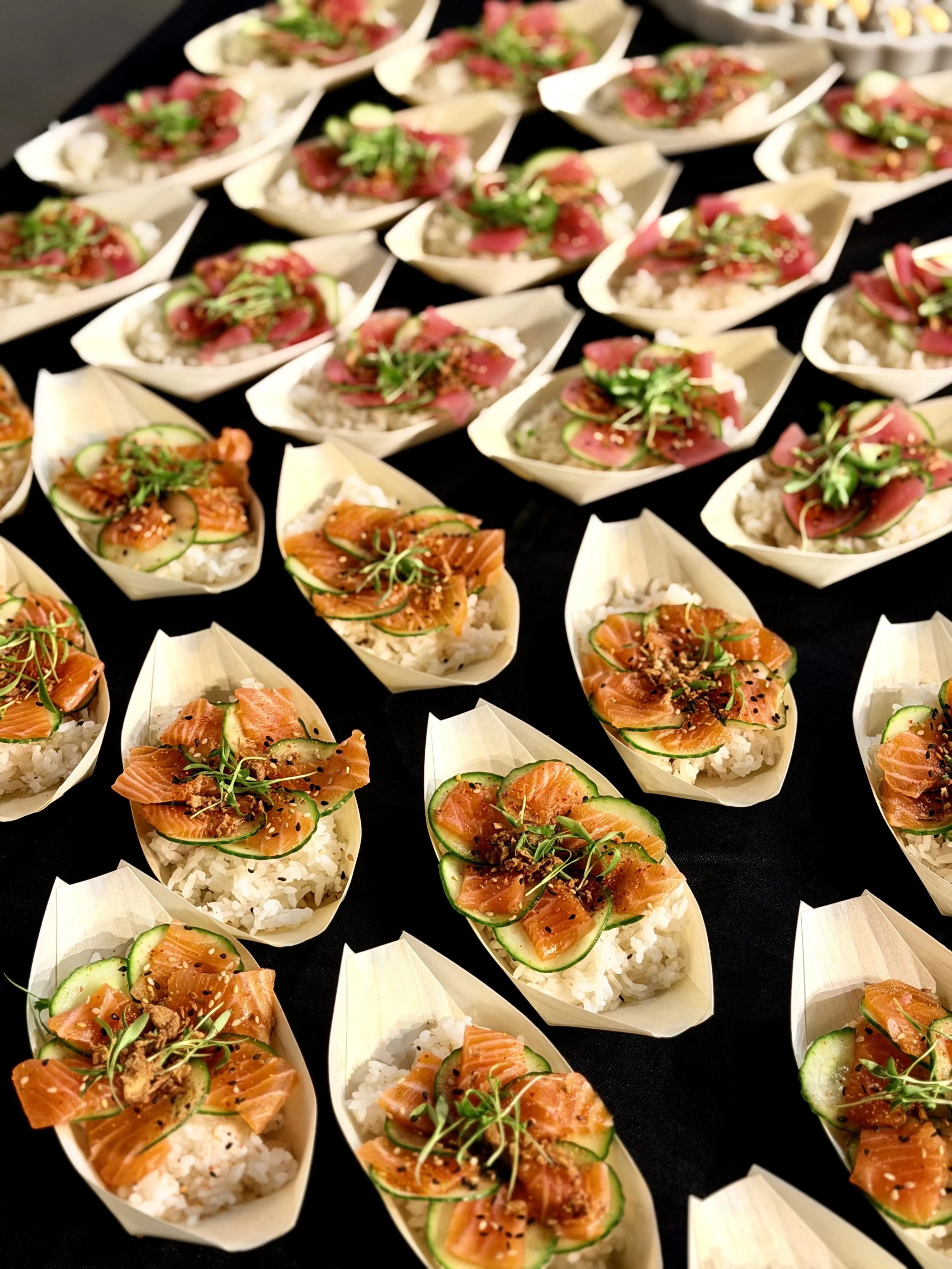 Multiple serving boats filled with Japanese-style sushi topped with raw fish, cucumber slices, and microgreens on a black surface.