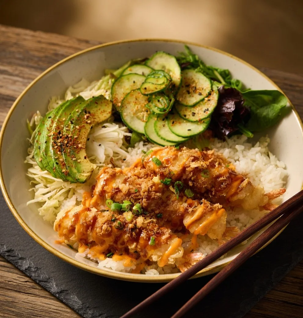 Bowl of Japanese tempura shrimp over rice with sliced cucumbers, avocado, and greens