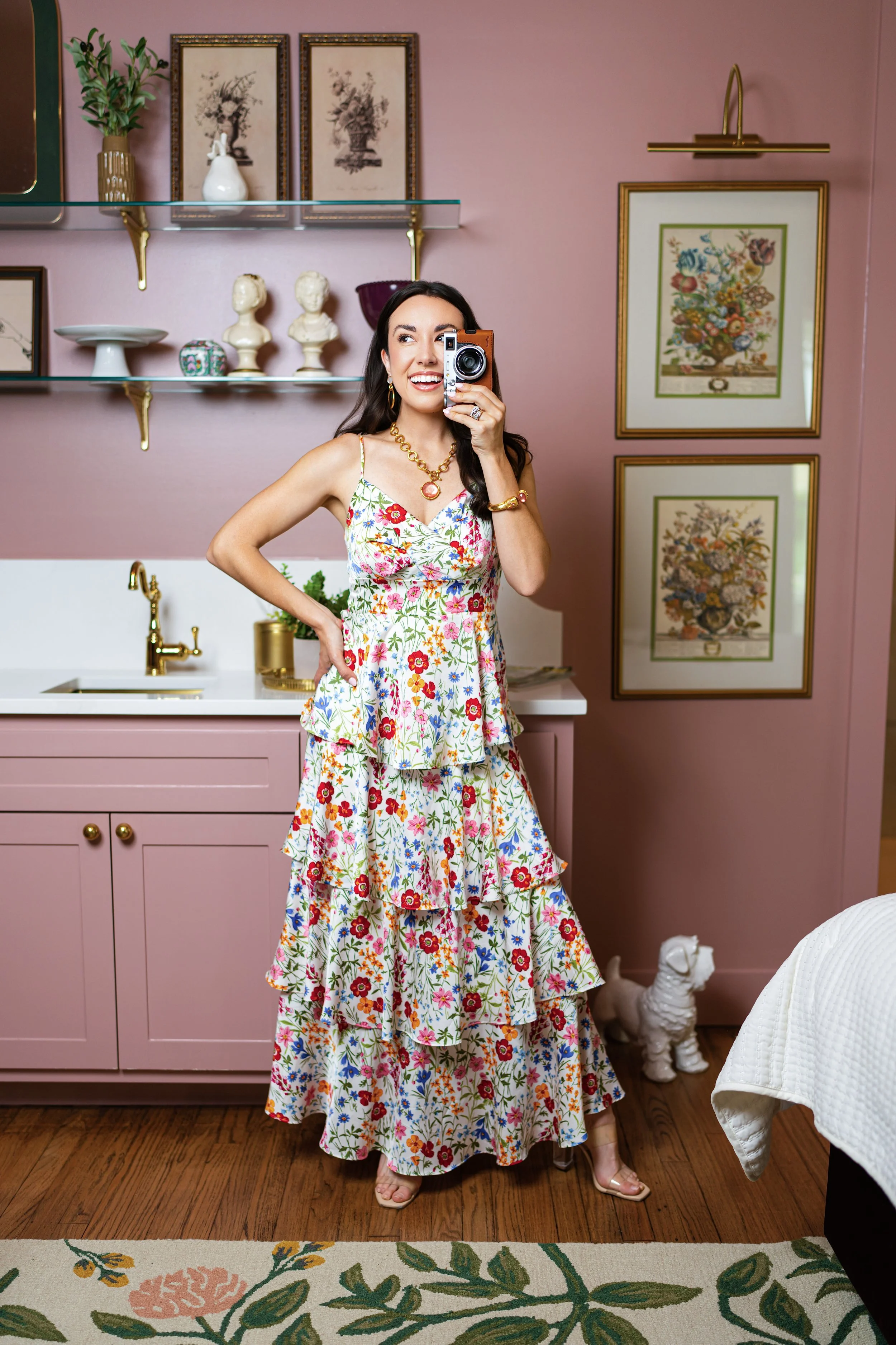 A woman with dark hair takes a selfie in a pink kitchen with gold accents. She wears a floral dress and gold jewelry. Behind her are framed botanical artwork, a pink wall, and shelves with decorative items. A dog statue is on the wooden floor near the kitchen cabinet.
