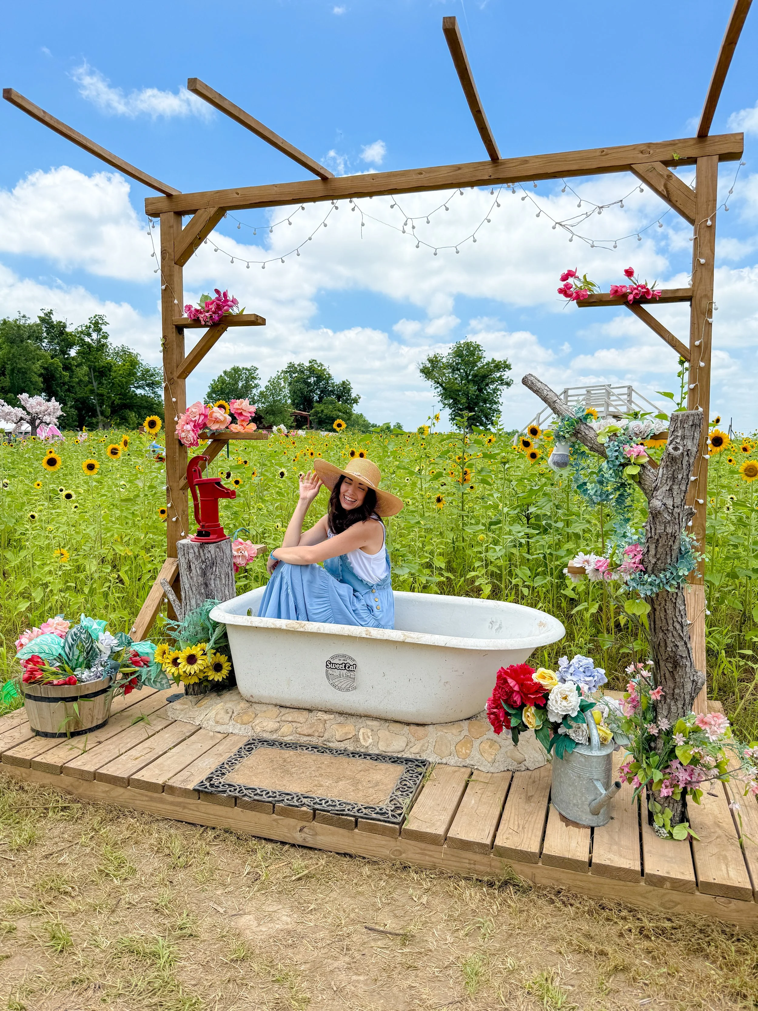 A woman sitting in a vintage bathtub outdoors, surrounded by flowers and greenery, with a wooden deck and an archway decorated with string lights and flowers. She is smiling, wearing a big sunhat, and raising her hand.
