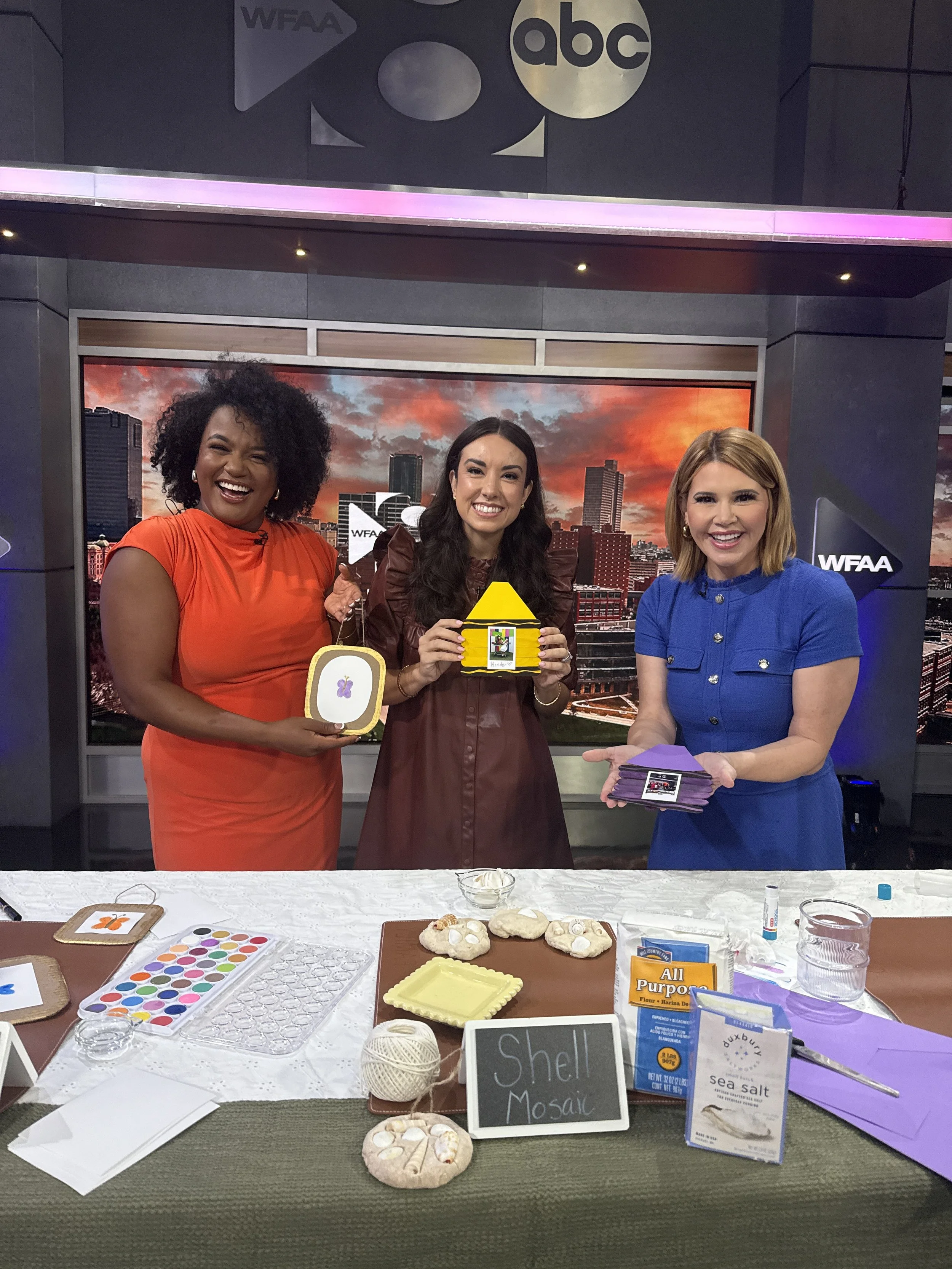 Three women standing behind a table displaying arts and crafts supplies, including painted rocks, colored paper, and decorative items, with a city skyline background on a screen and signs indicating craft projects.