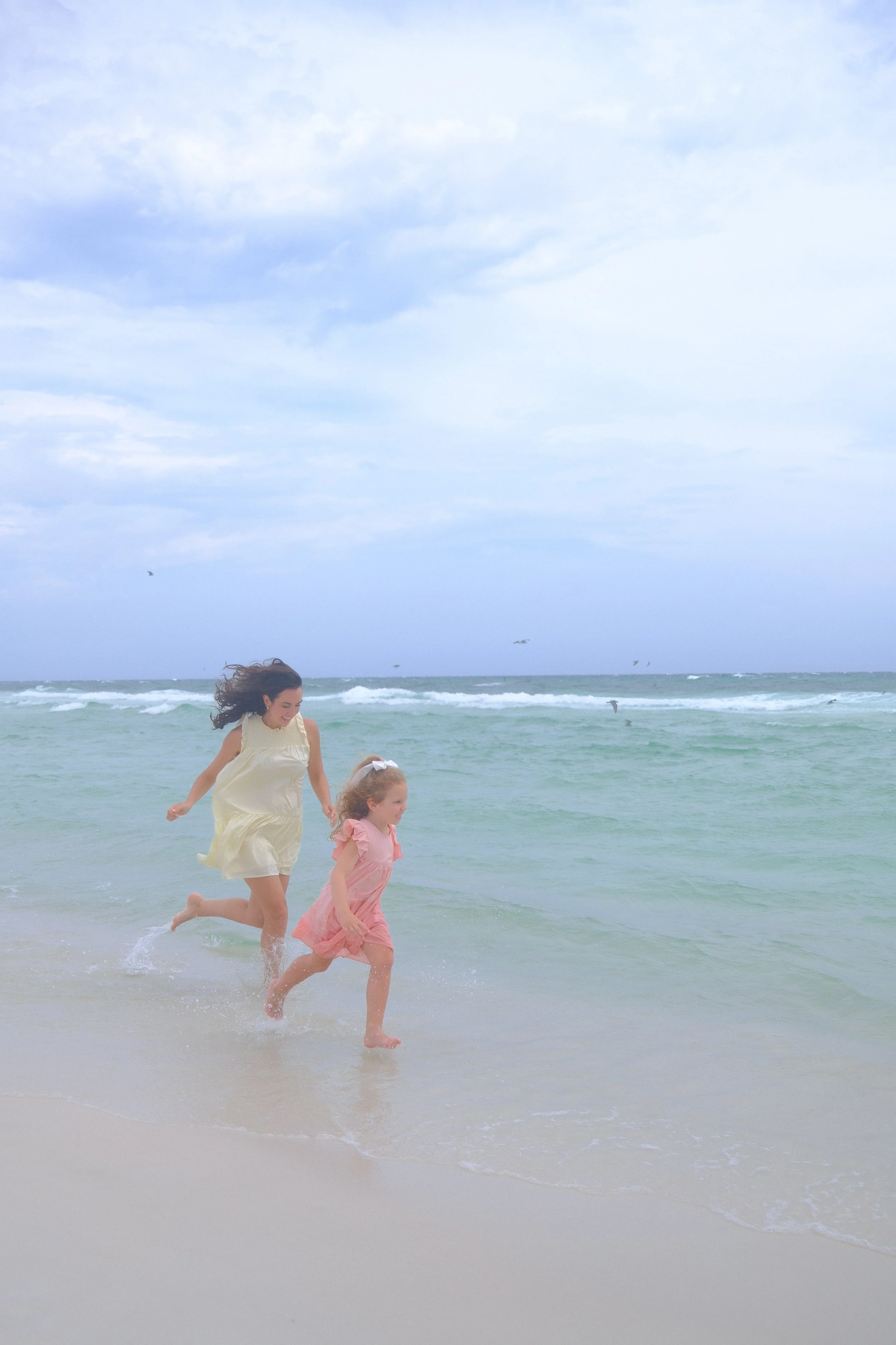 Two young girls, one in a cream dress and the other in a pink dress, running and playing at the beach near the water's edge, with waves and a cloudy sky in the background.