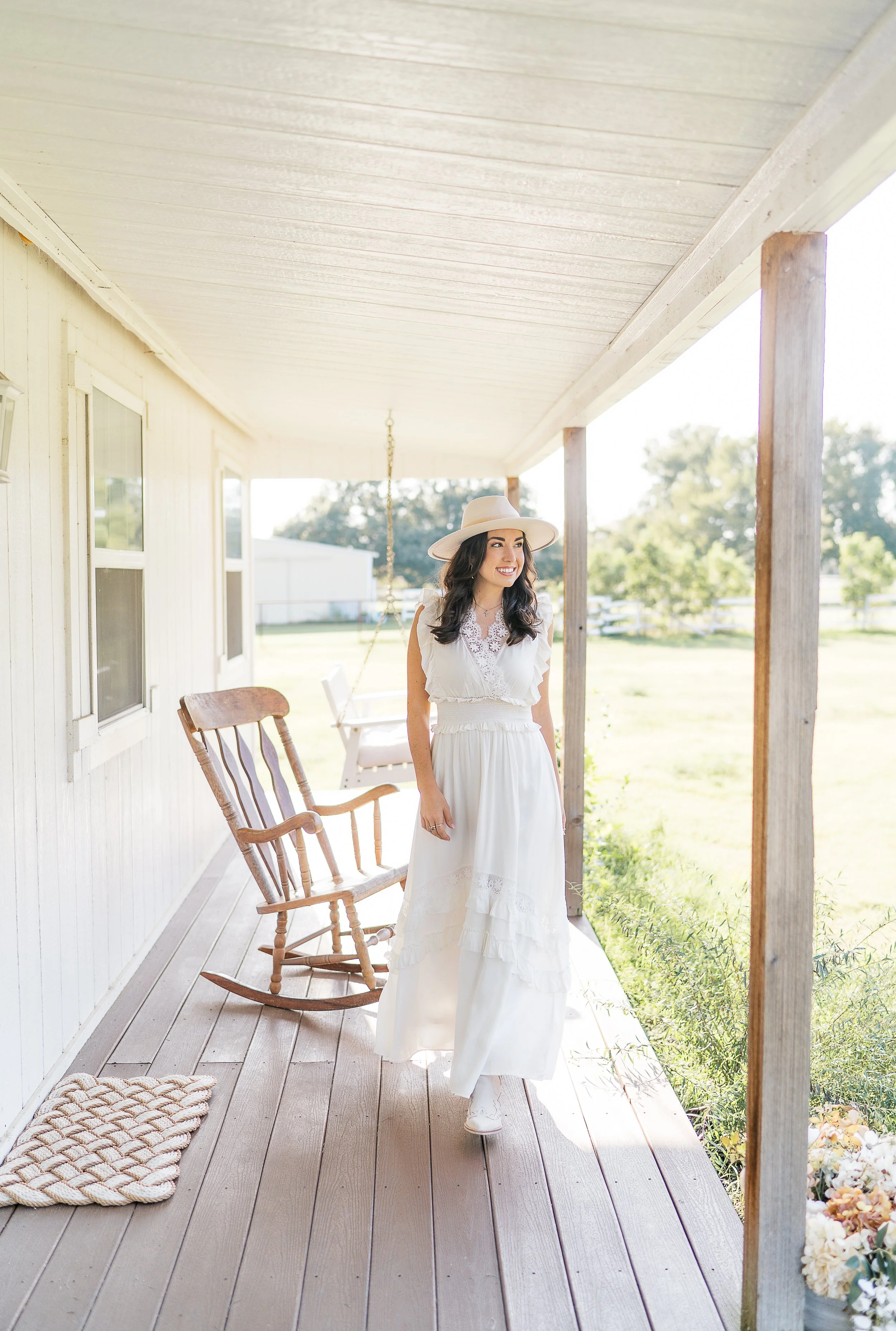 A woman in a white dress and large white hat stands on a porch, smiling, with rocking chairs and greenery in the background.