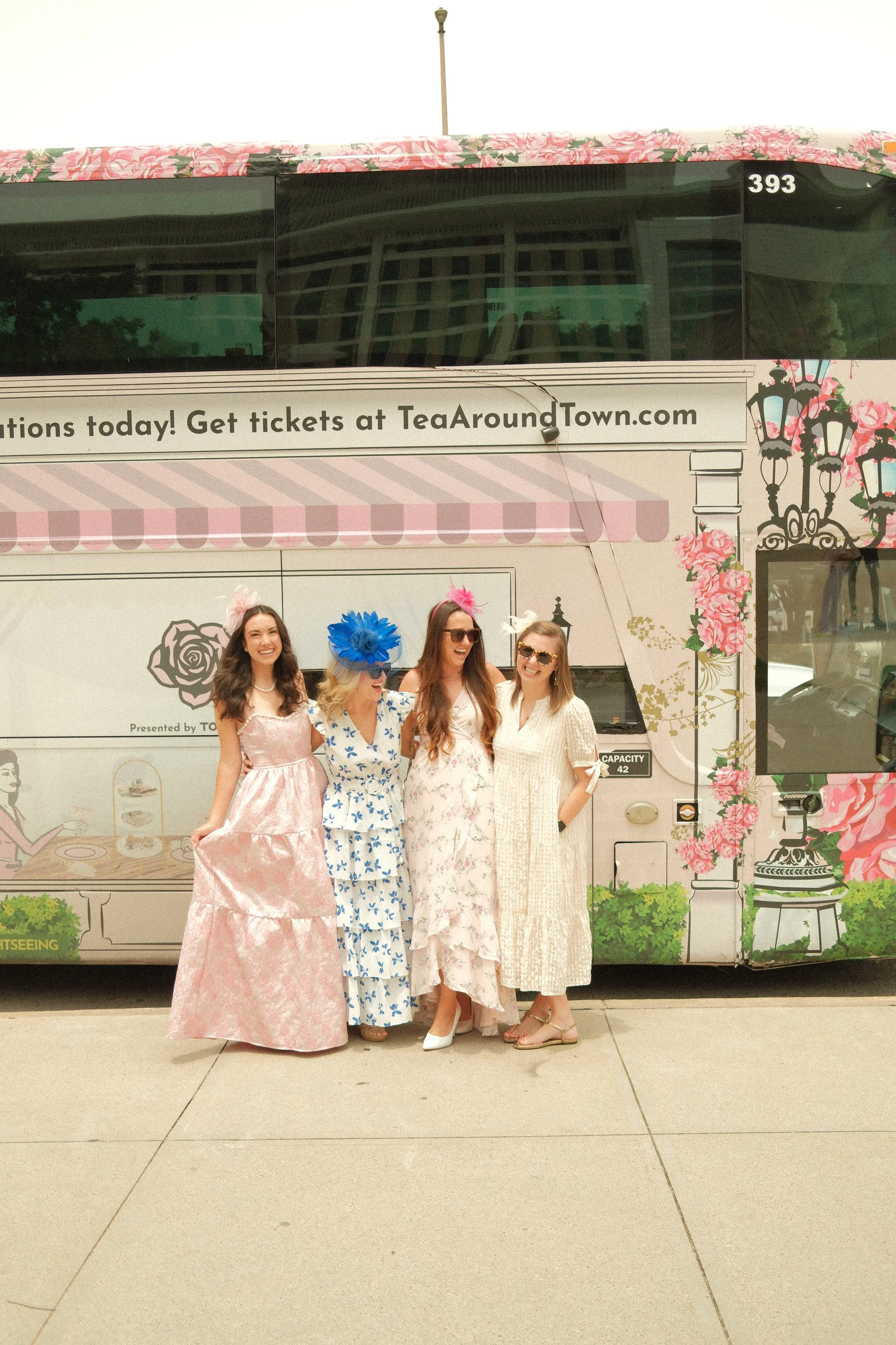 Four women in vintage-style dresses standing in front of a decorated tea-themed vehicle with pink floral and street lamp motifs. They are outdoors on a sidewalk.