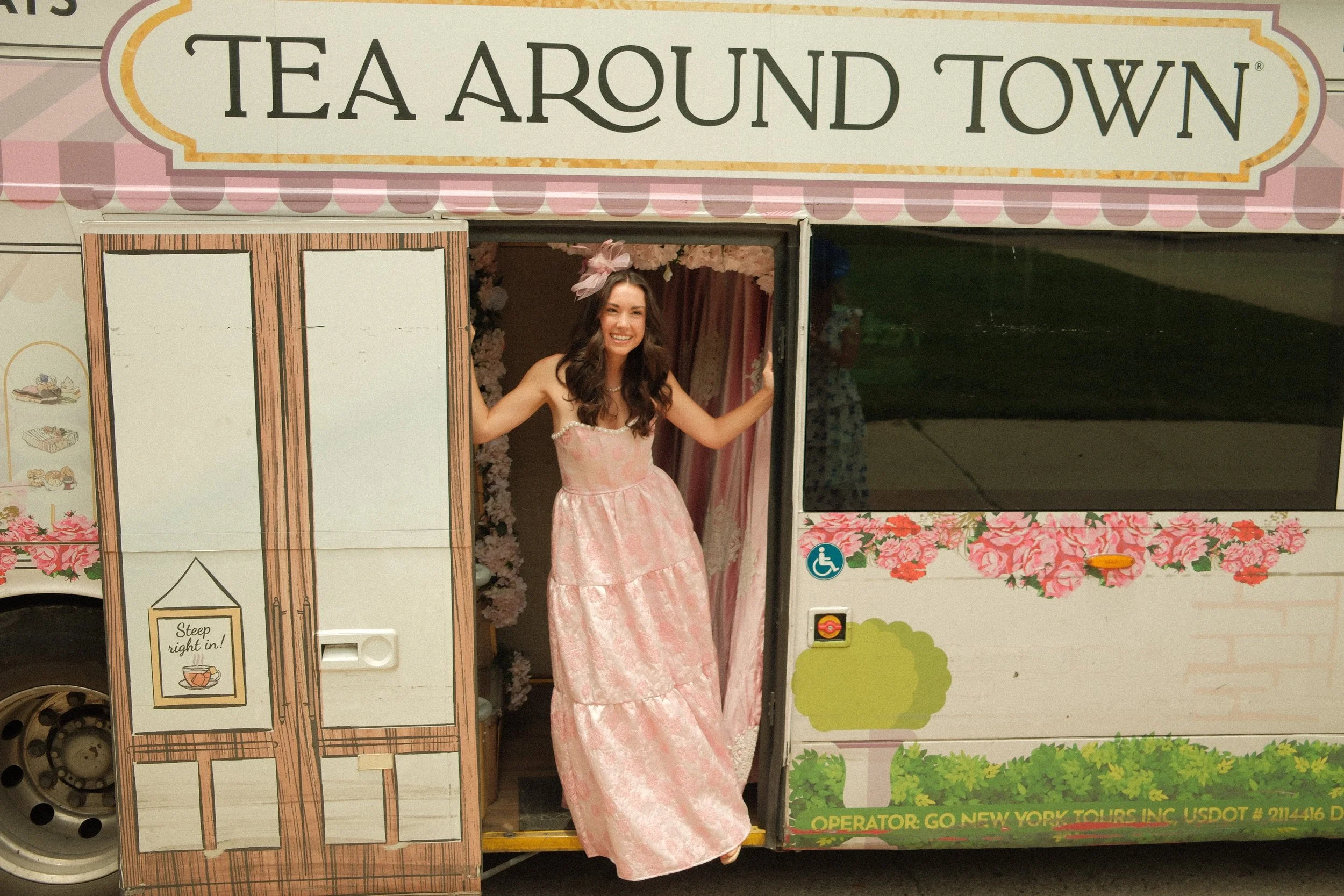 A young woman in a pink dress with a large pink bow in her hair, smiling and standing in the doorway of a themed food truck called 'Tea Around Town.' The truck has floral and tea-themed decorations, and a sign that says 'Sleep tight in!'