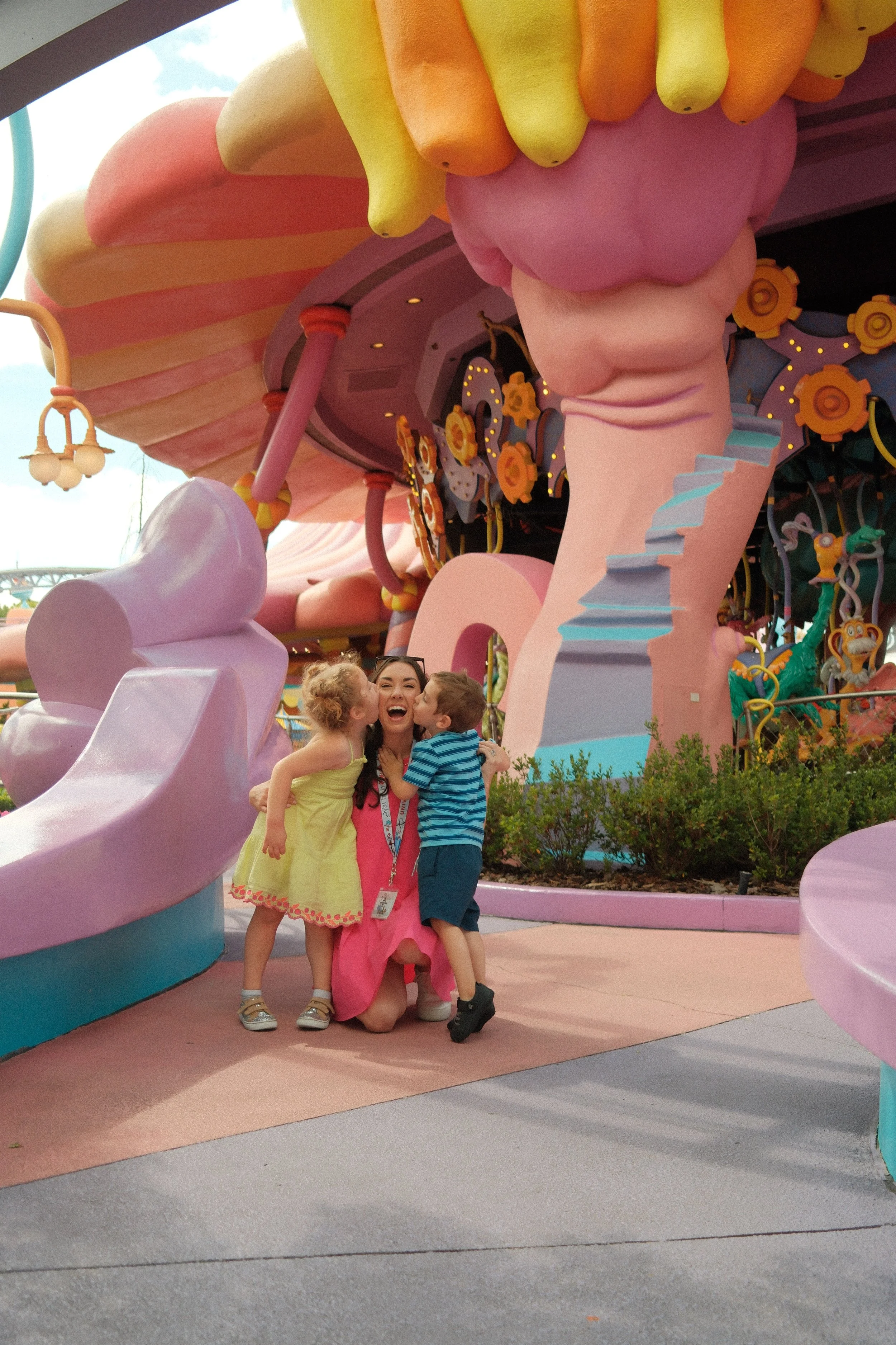 A woman in a pink dress is kneeling on the ground, hugging and kissing two children, a girl in a yellow dress and a boy in a striped shirt, at an animated themed area with pastel-colored structures and whimsical decorations.