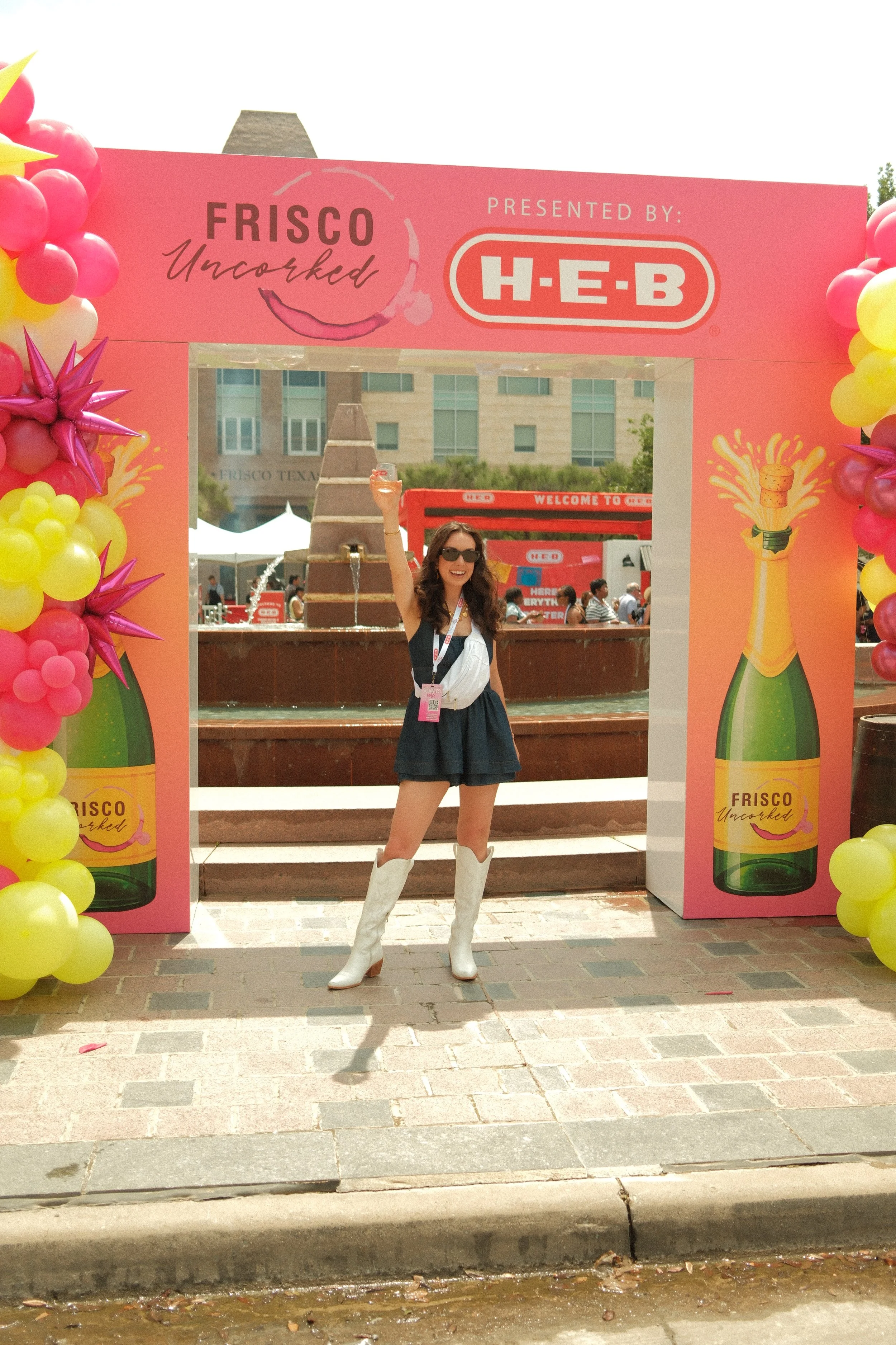 A young woman wearing sunglasses, a black skirt, white cowboy boots, and a white jacket standing under a pink and yellow balloon arch during an outdoor event, smiling and raising her arm.