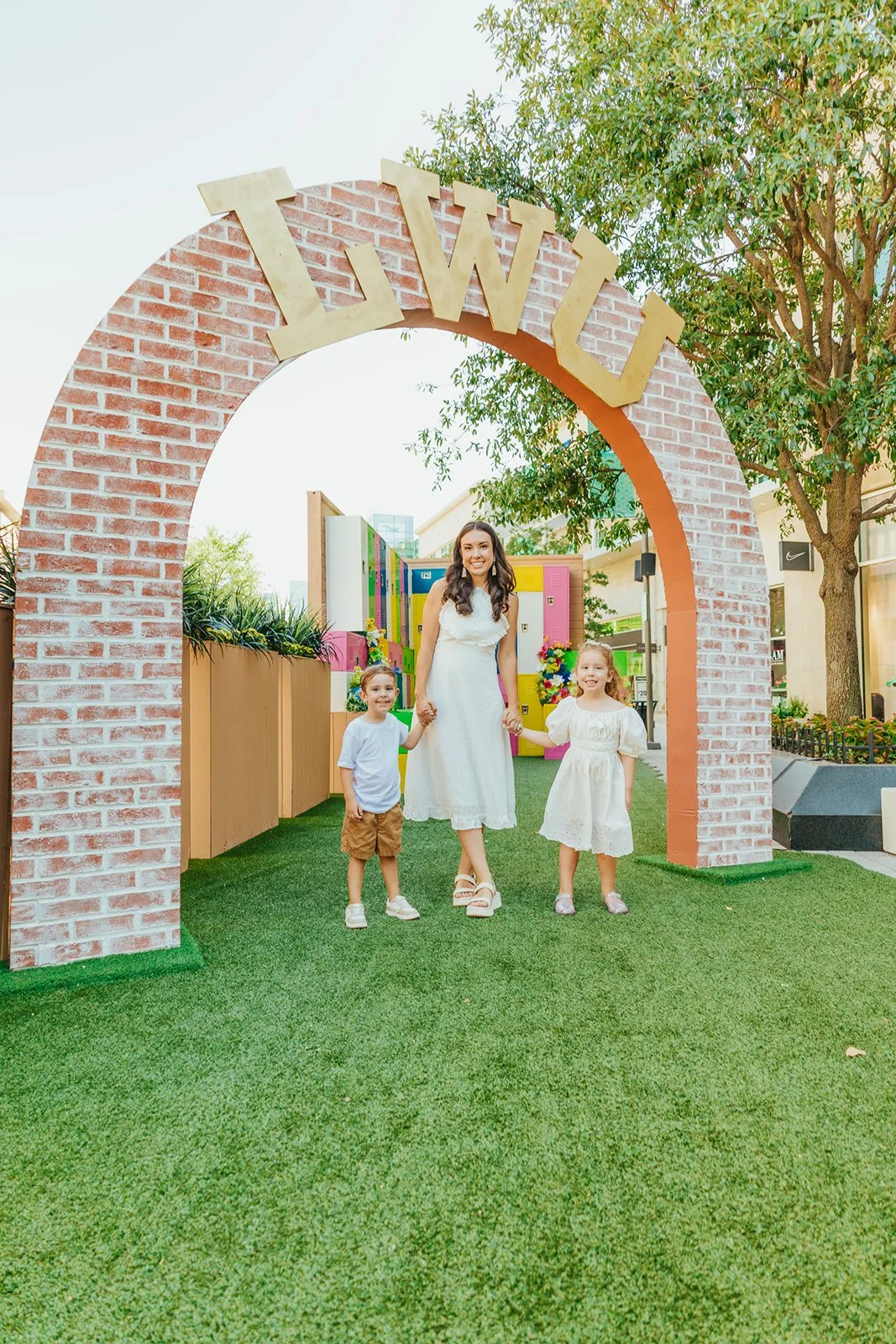A woman and two children holding hands under a brick arch with the word 'WILLI' on top, in an outdoor setting with trees, colorful lockers, and green grass.