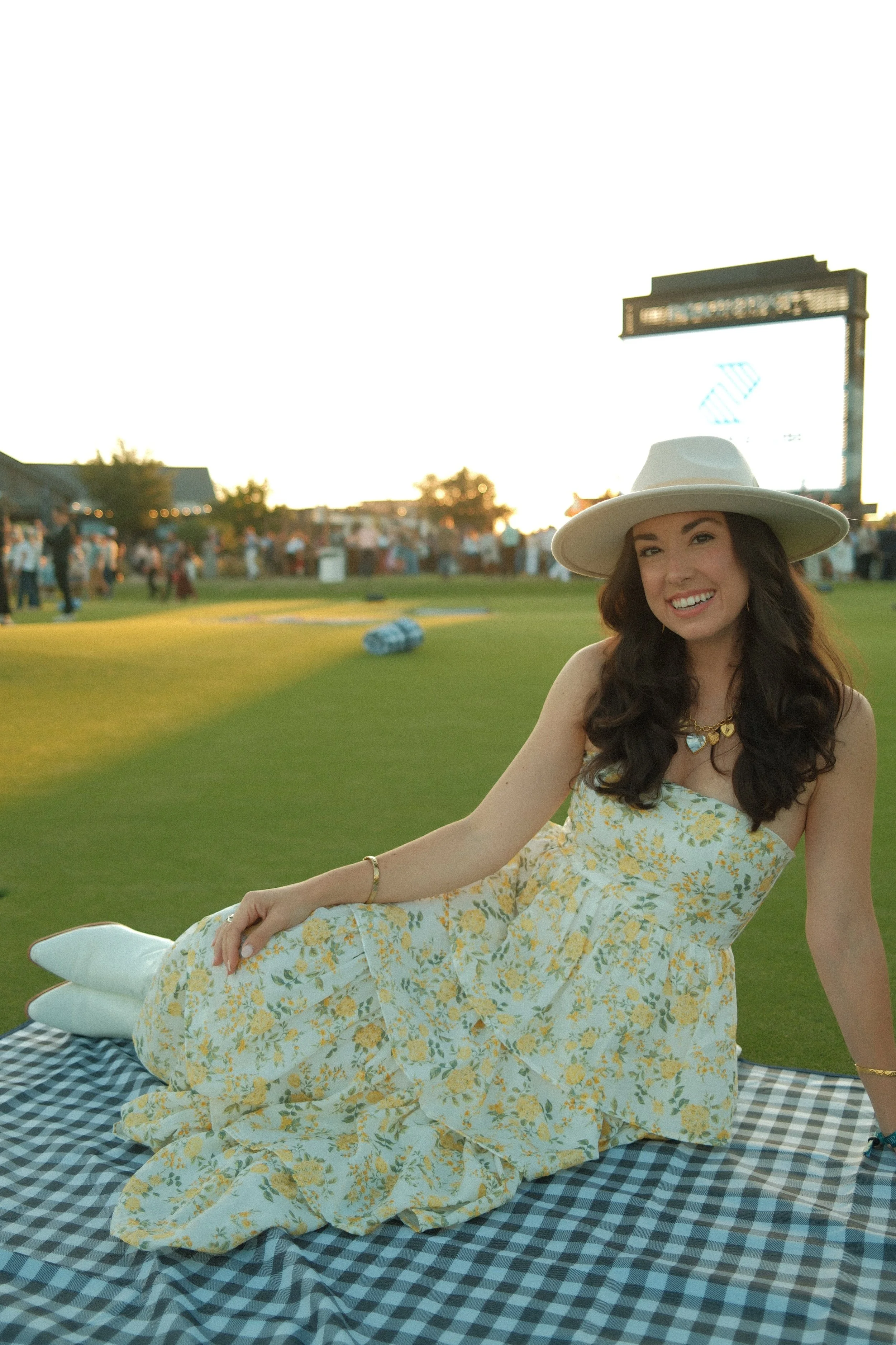 A smiling woman in a floral dress and wide-brimmed hat sitting on a checkered blanket at a park during sunset, with a crowd and an electronic billboard in the background.