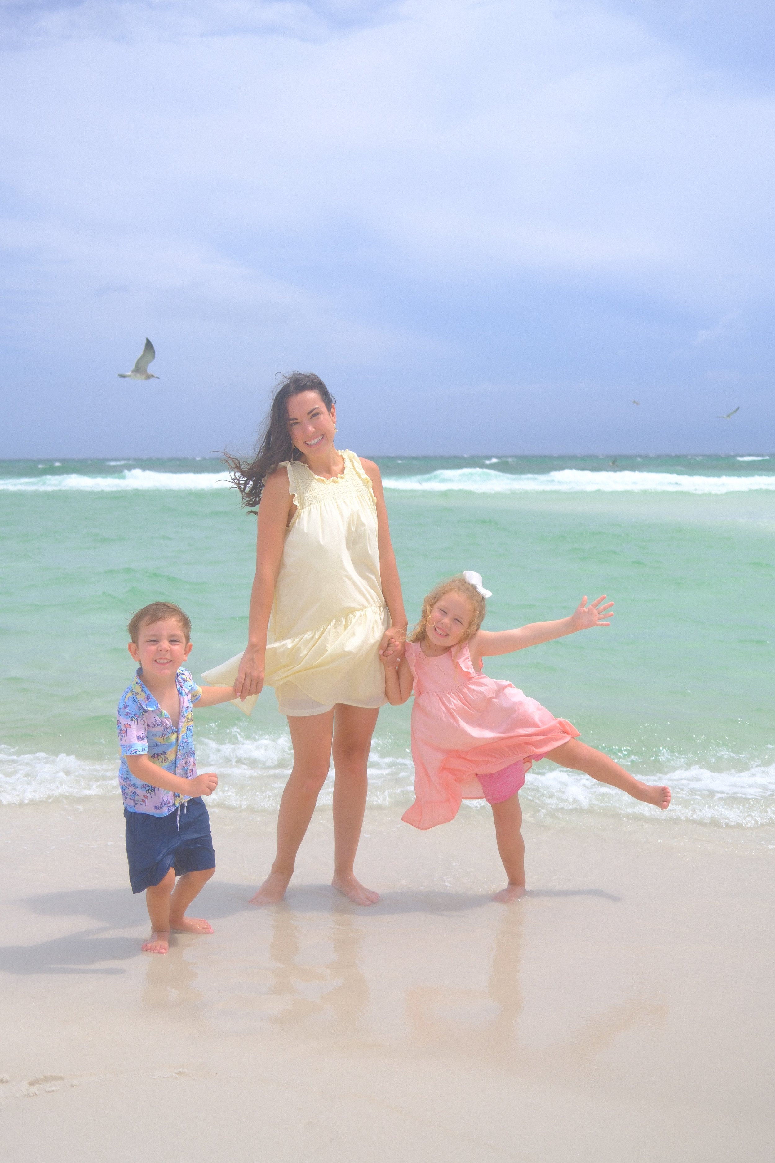 A woman and three children playing on a beach, with the woman in a cream dress and the children in colorful outfits, and seagulls flying in the cloudy sky.