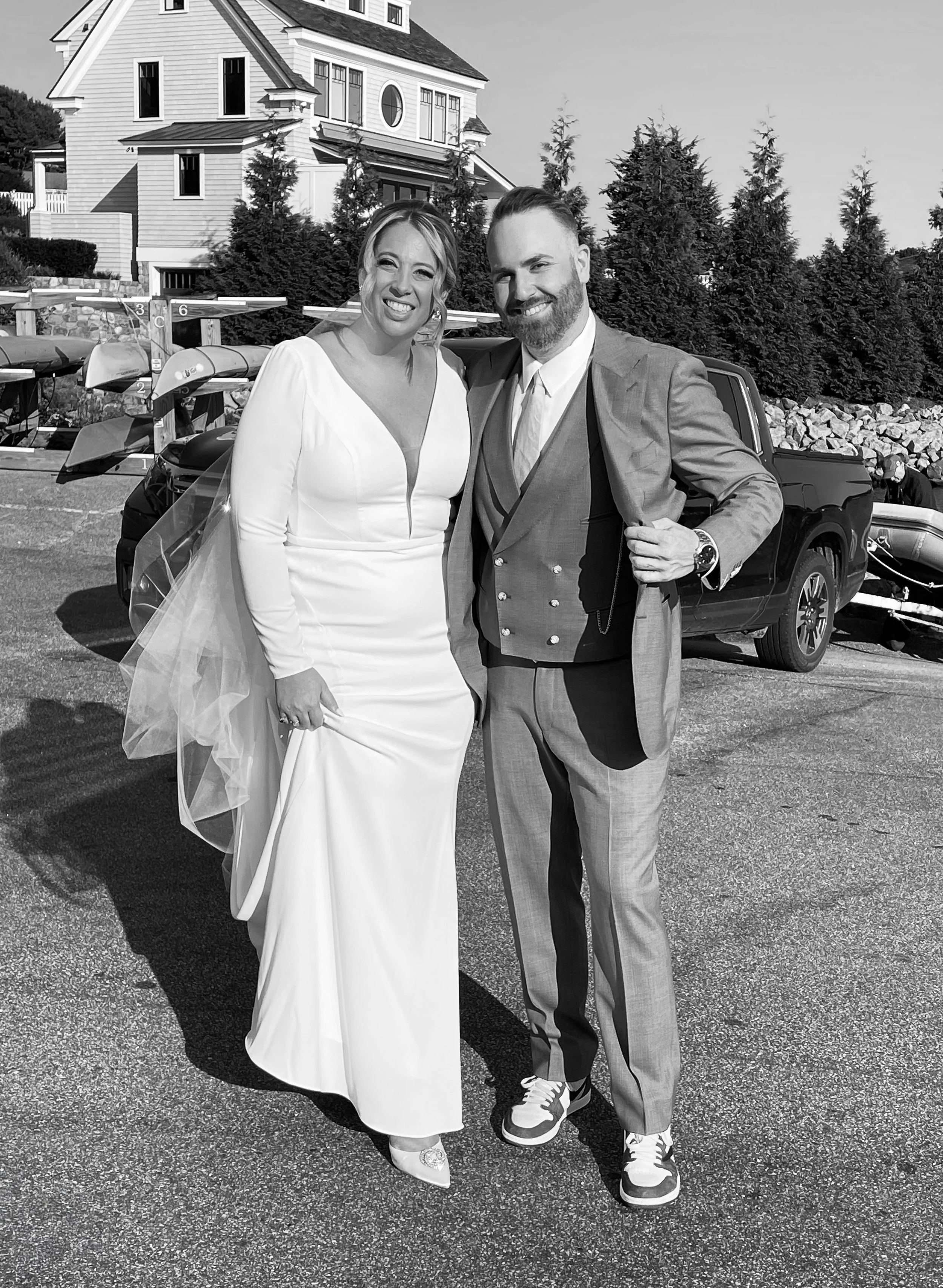A bride and groom posing together outside on a paved area, with kayaks and a house in the background. The bride is wearing a long white wedding gown with a deep V neckline and veil, and the groom is in a gray suit with a vest, tie, and sneakers, smiling at the camera.