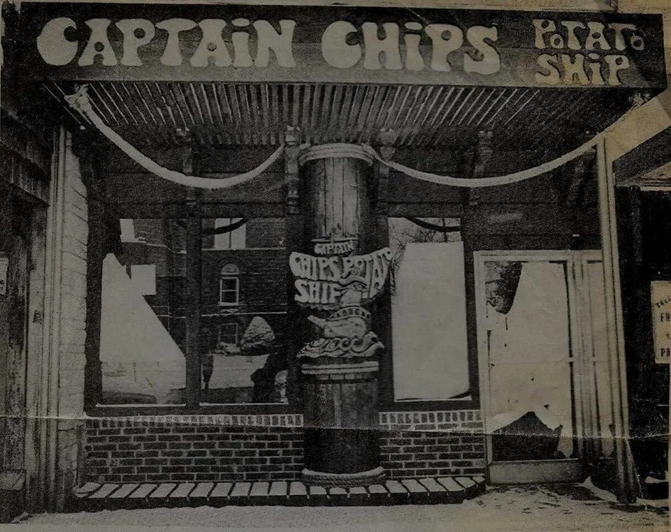 Black and white photo of a storefront with a sign that reads 'Captain Chips' and 'Fried Shrimp'. The storefront features brickwork and large windows, with a decorative pillar in the center.