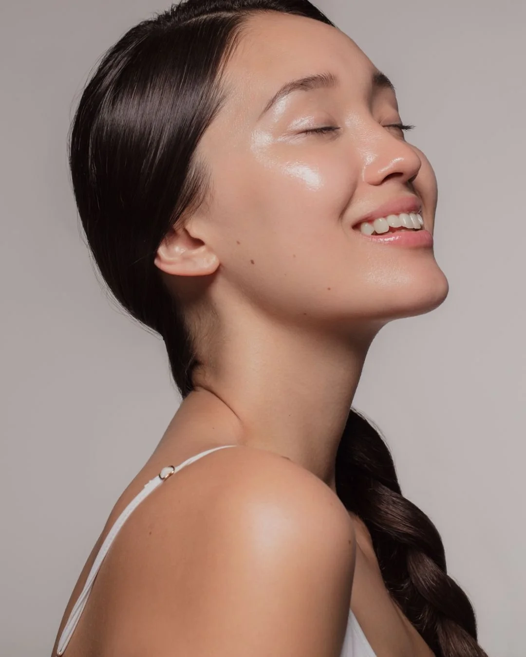 Close-up of a woman with long, dark hair styled in a braid, smiling with closed eyes, showing natural makeup and glowing skin, against a neutral background.