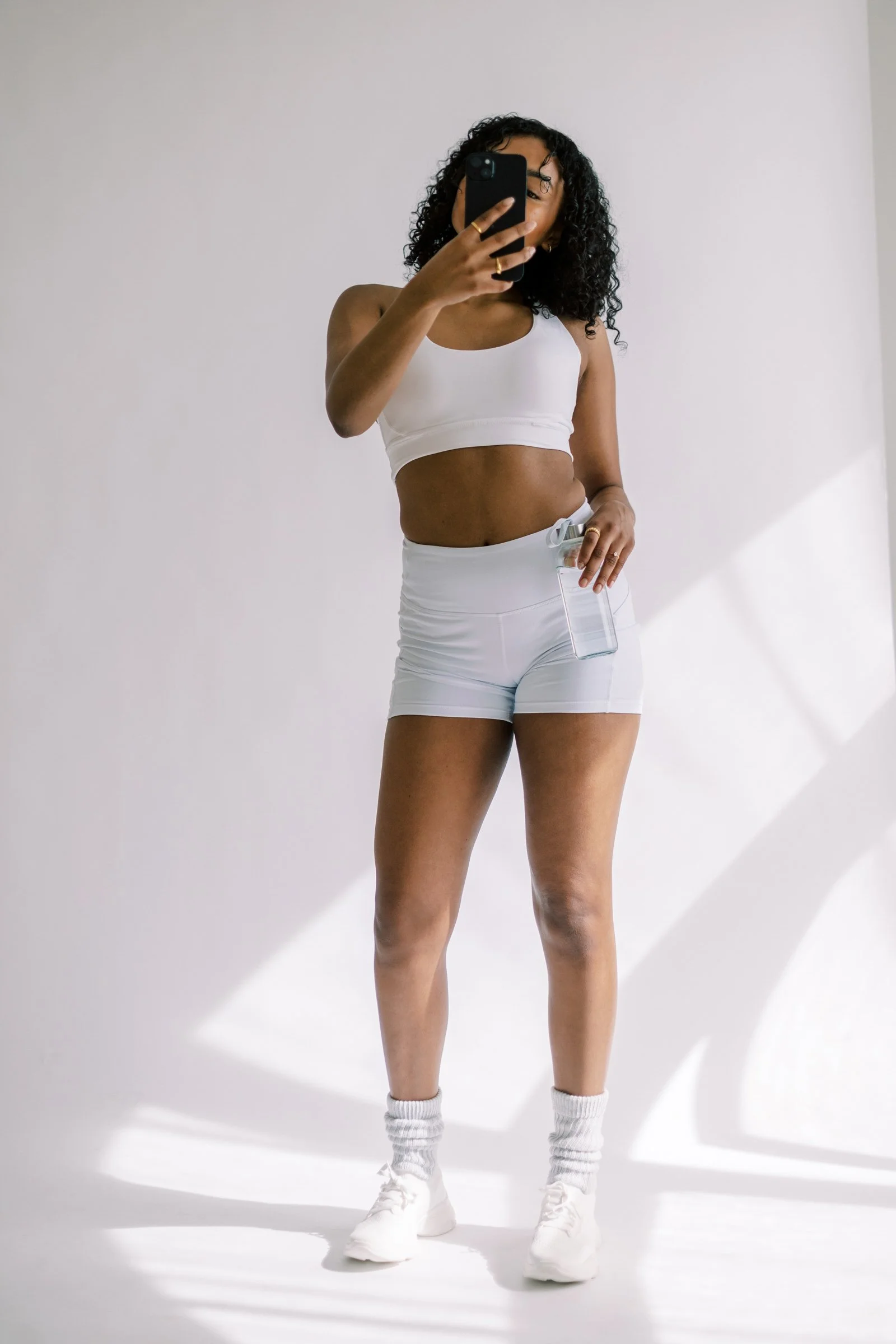 A woman taking a mirror selfie in athletic wear, wearing a white sports bra and shorts, white sneakers, and holding a fitness water bottle.