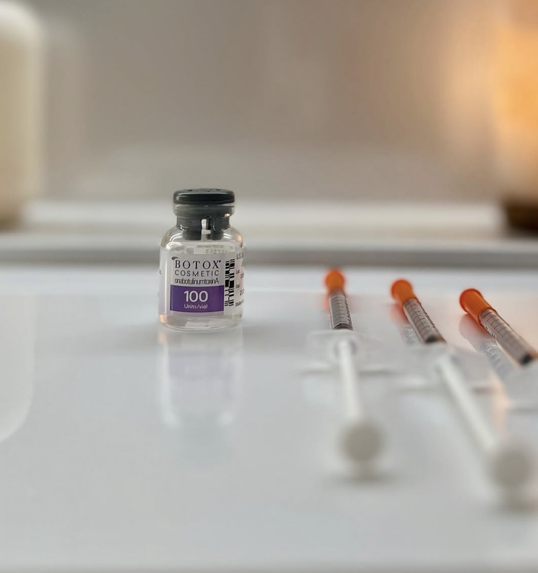 A small vial labeled 'Botox Cosmetic' with 100 units, placed on a white surface, with three syringes lying nearby, all ready for a cosmetic procedure.