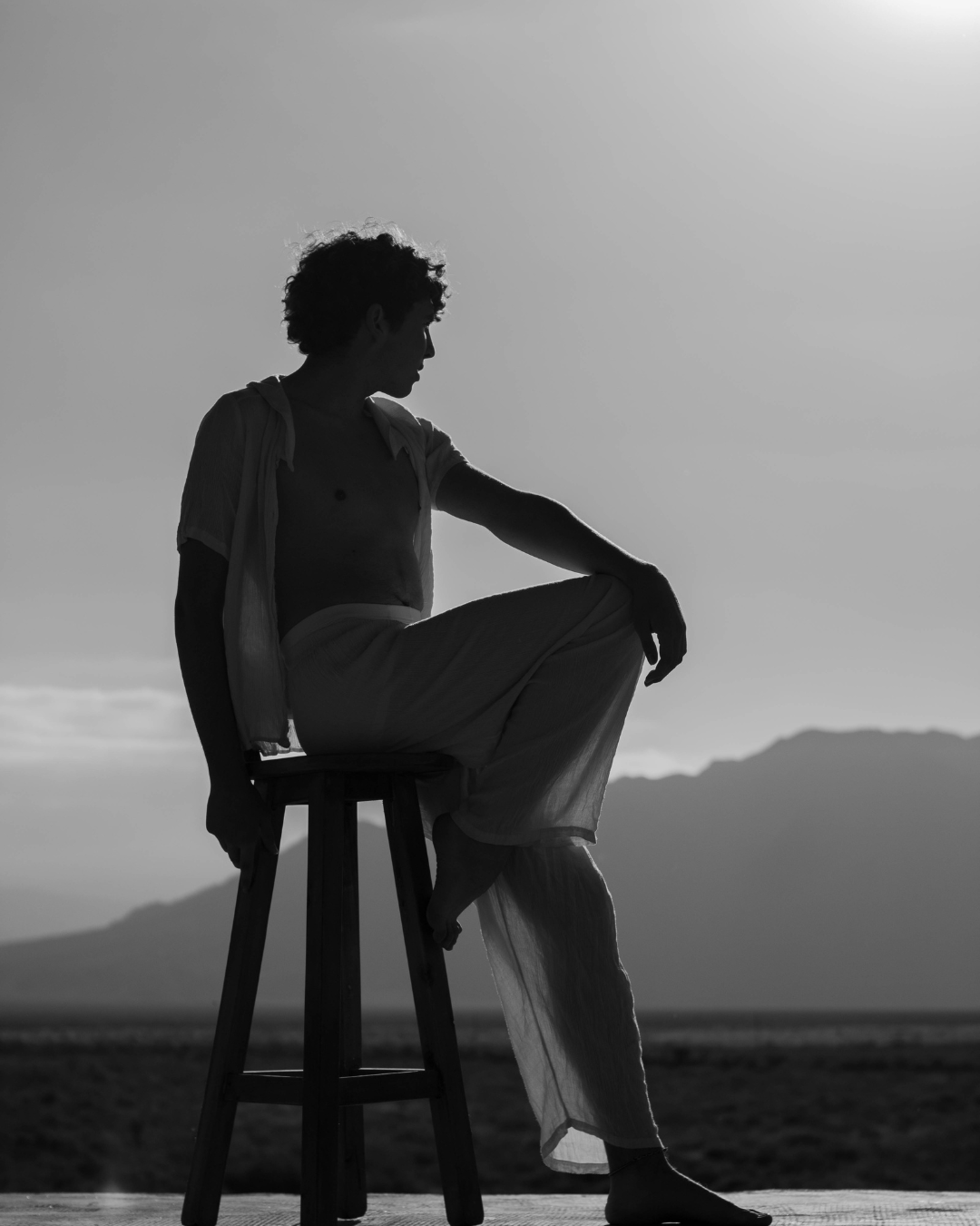 Silhouette of a woman sitting on a stool outdoors, with mountains in the background, captured in black and white.