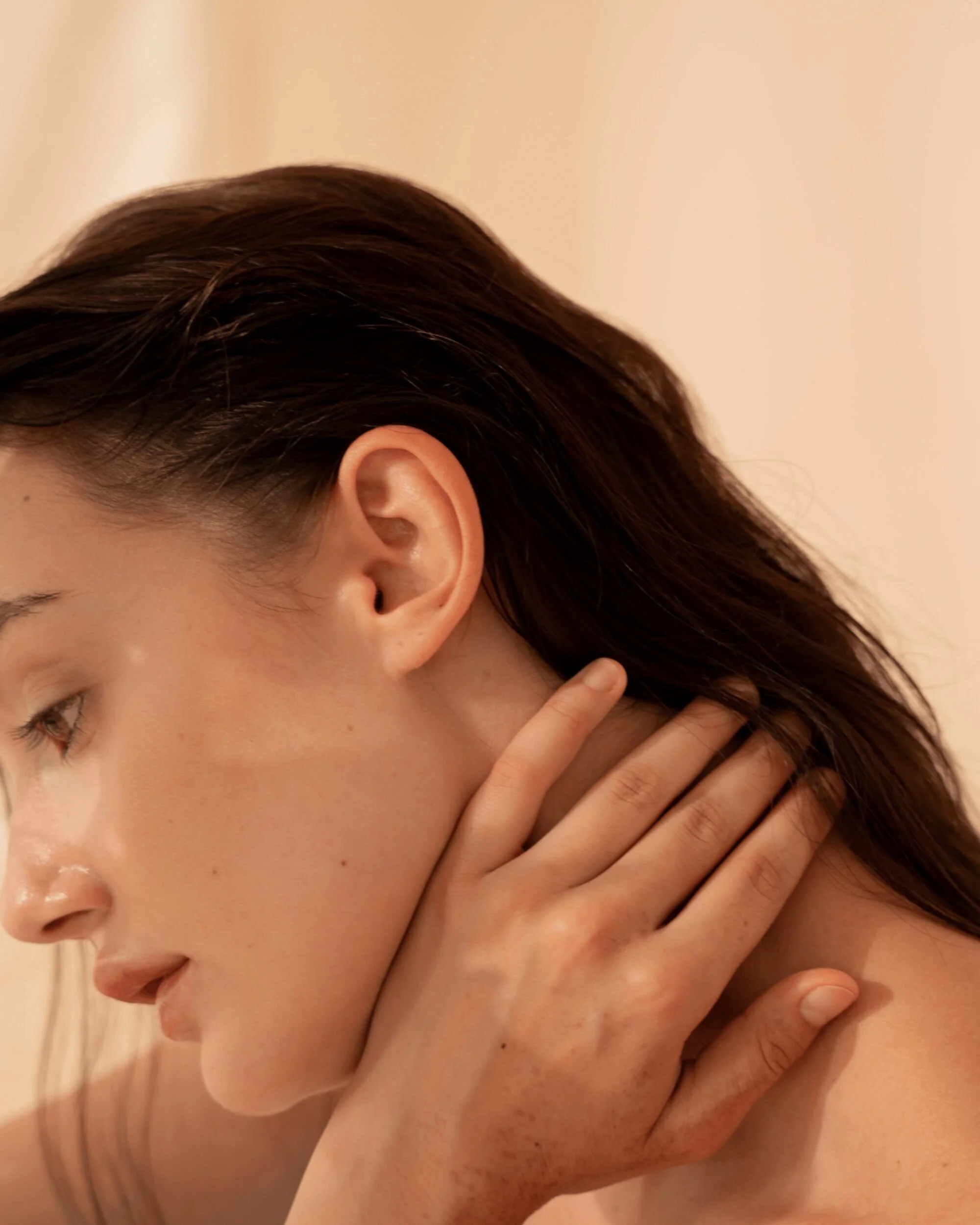 A young woman with dark brown hair and fair skin, touching her neck with her left hand, seen in a close-up profile shot against a neutral background.
