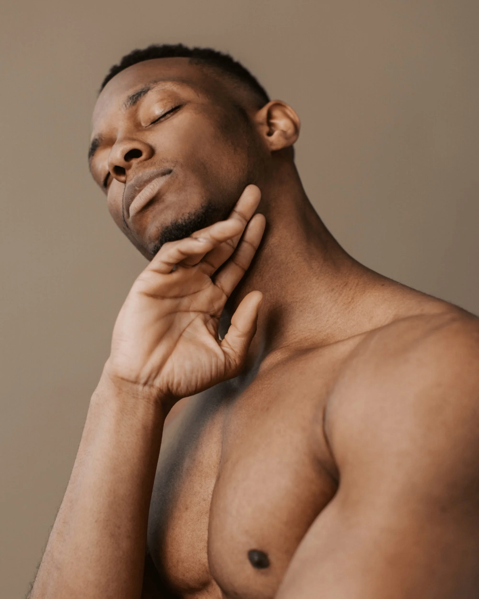 A shirtless Black man with closed eyes and a relaxed expression touches his neck with one hand.