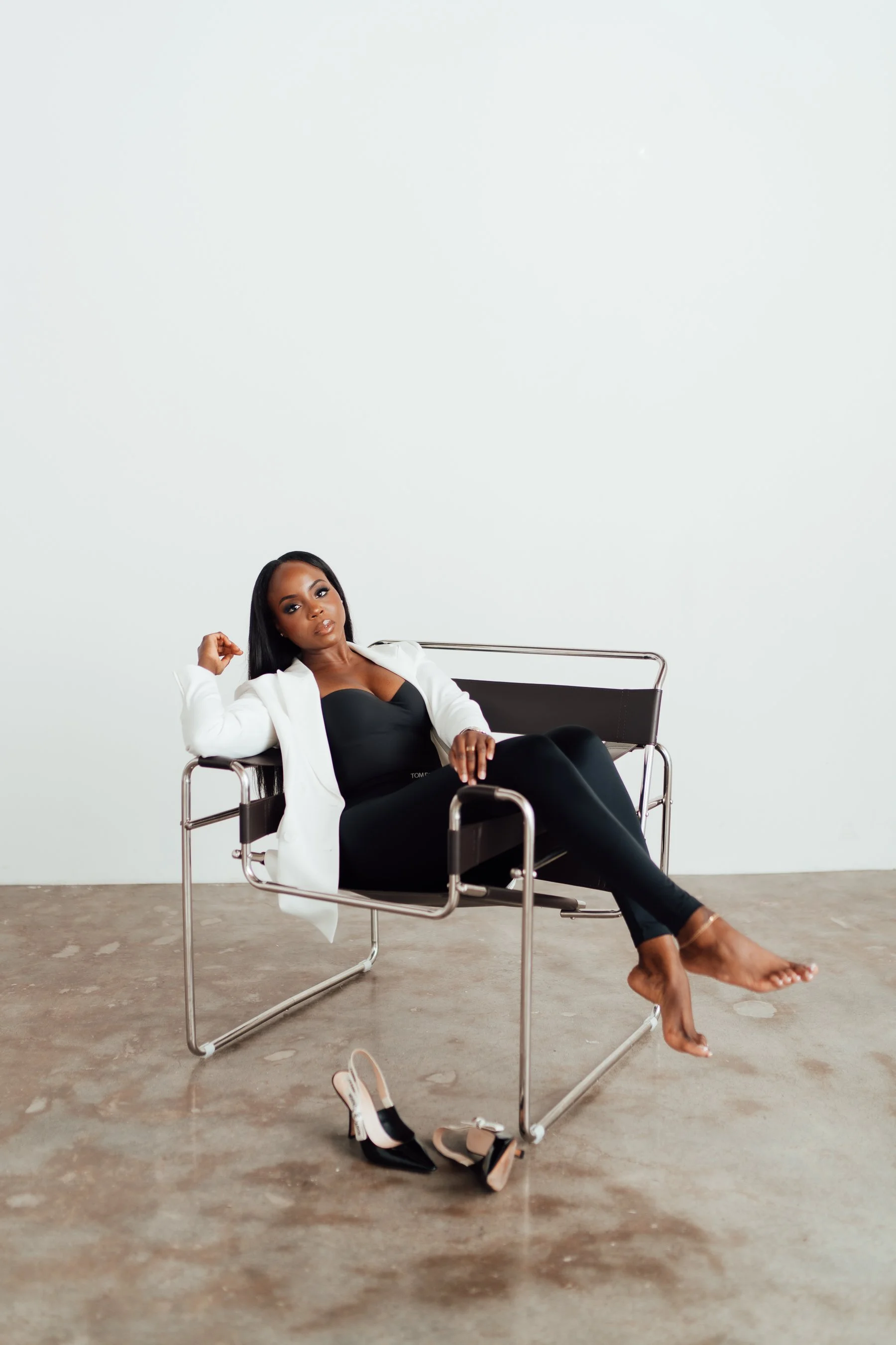 Woman in a black dress and white blazer sitting barefoot on a black chair, with a pair of high heels on the floor in front of her, in a minimalist room with a concrete floor and white wall.