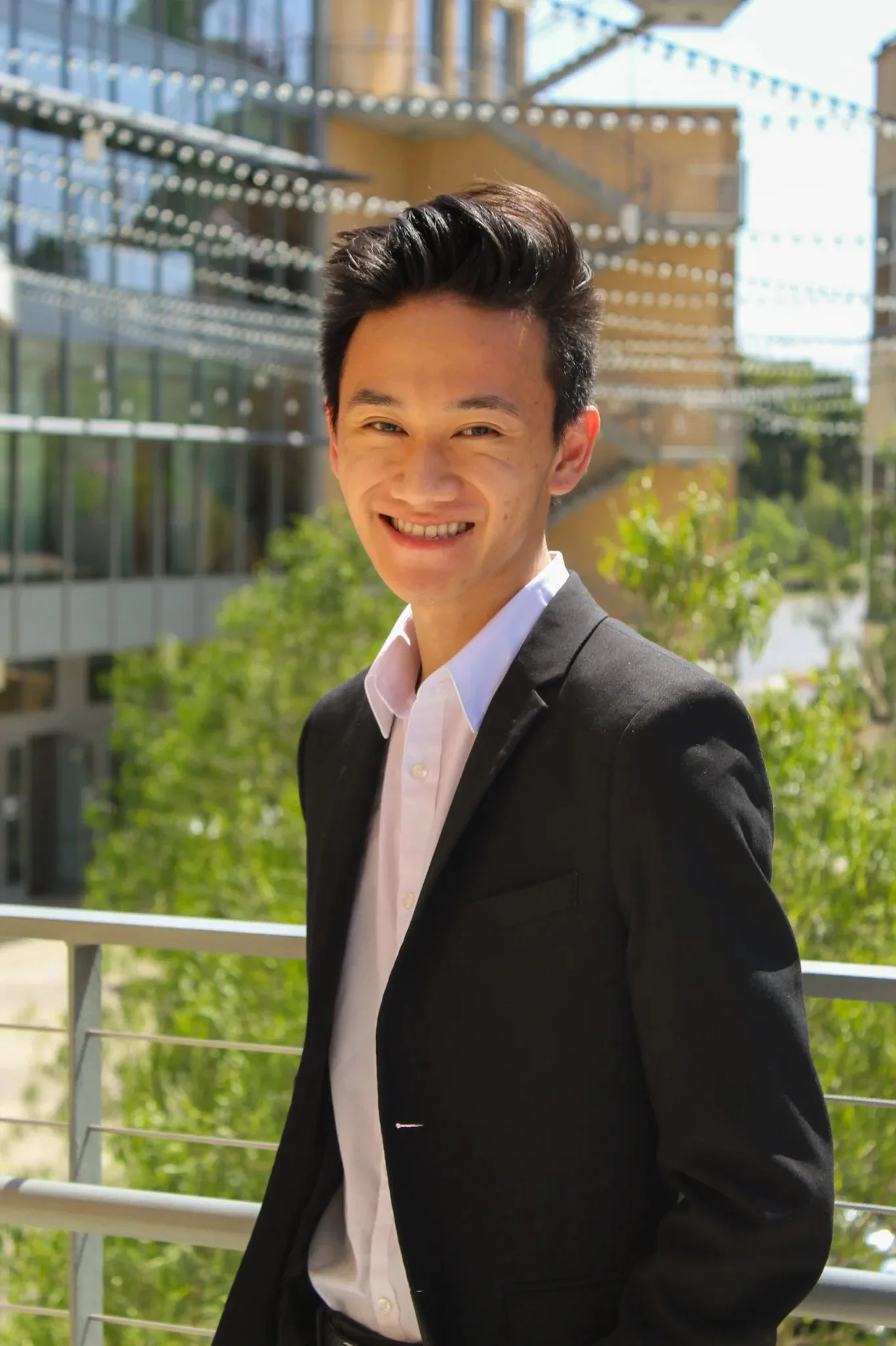 Person in a suit smiling outdoors in front of a modern building with glass windows and greenery in the background.
