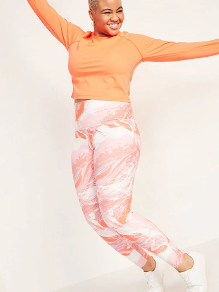 Woman smiling, jumping, wearing orange long-sleeve top and pink and white patterned leggings.