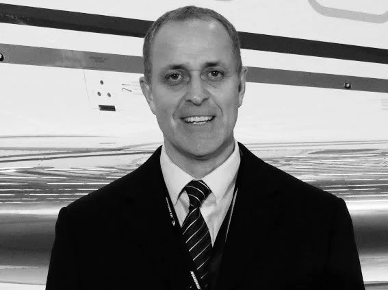 A man in a suit wearing a striped tie, smiling, with a lanyard around his neck, standing in front of a modern metallic wall.