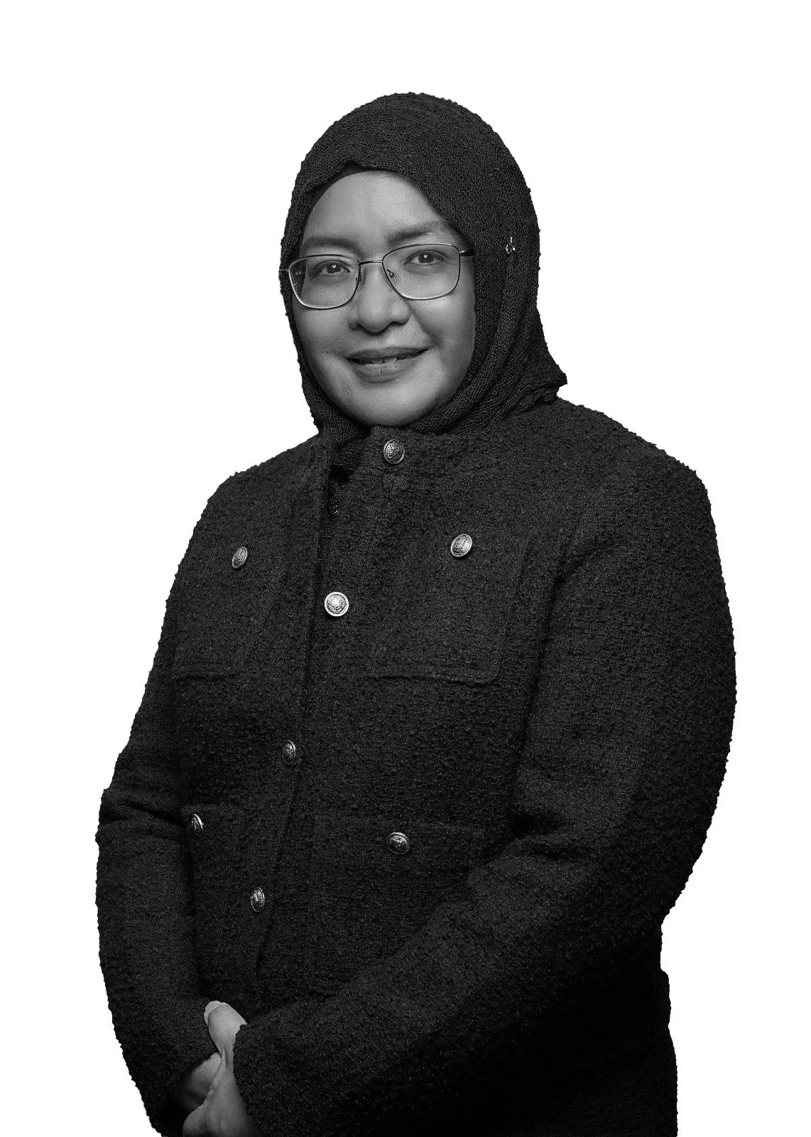 Black and white portrait of a woman wearing glasses, a hijab, and a textured coat with buttons, standing against a plain background.