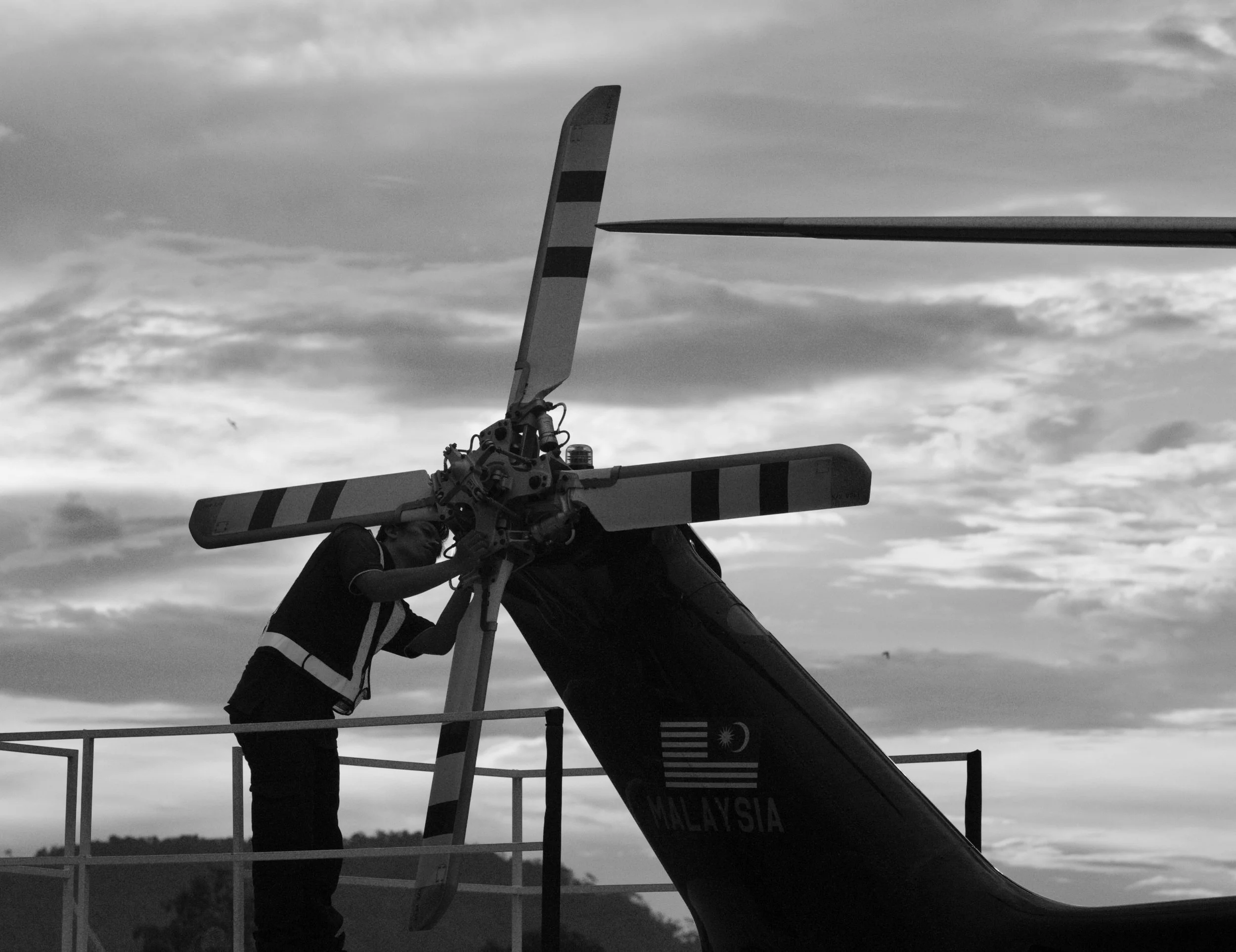 A person in a safety vest working on a helicopter rotor on an outdoor tarmac at sunset, with a cloudy sky in the background.