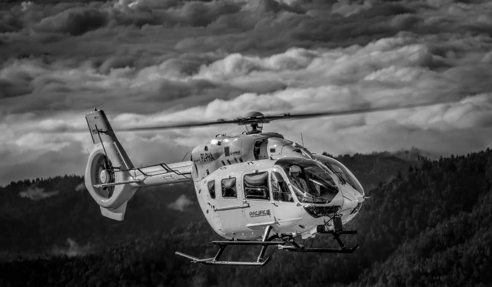 Black and white image of a helicopter flying over a mountainous landscape with cloudy skies in the background.
