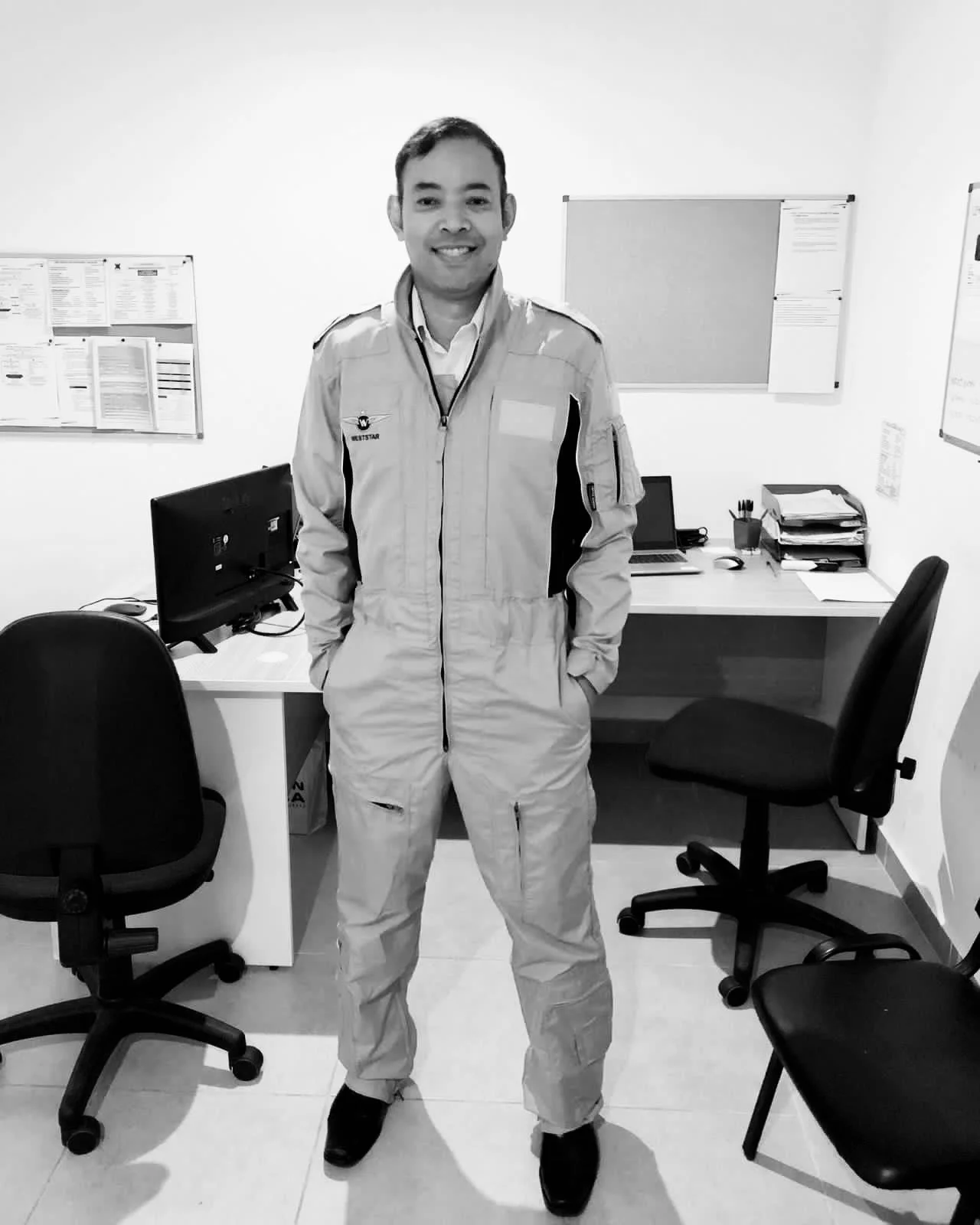 Black and white photo of a man in a flight suit standing in an office with desks, chairs, and computers.