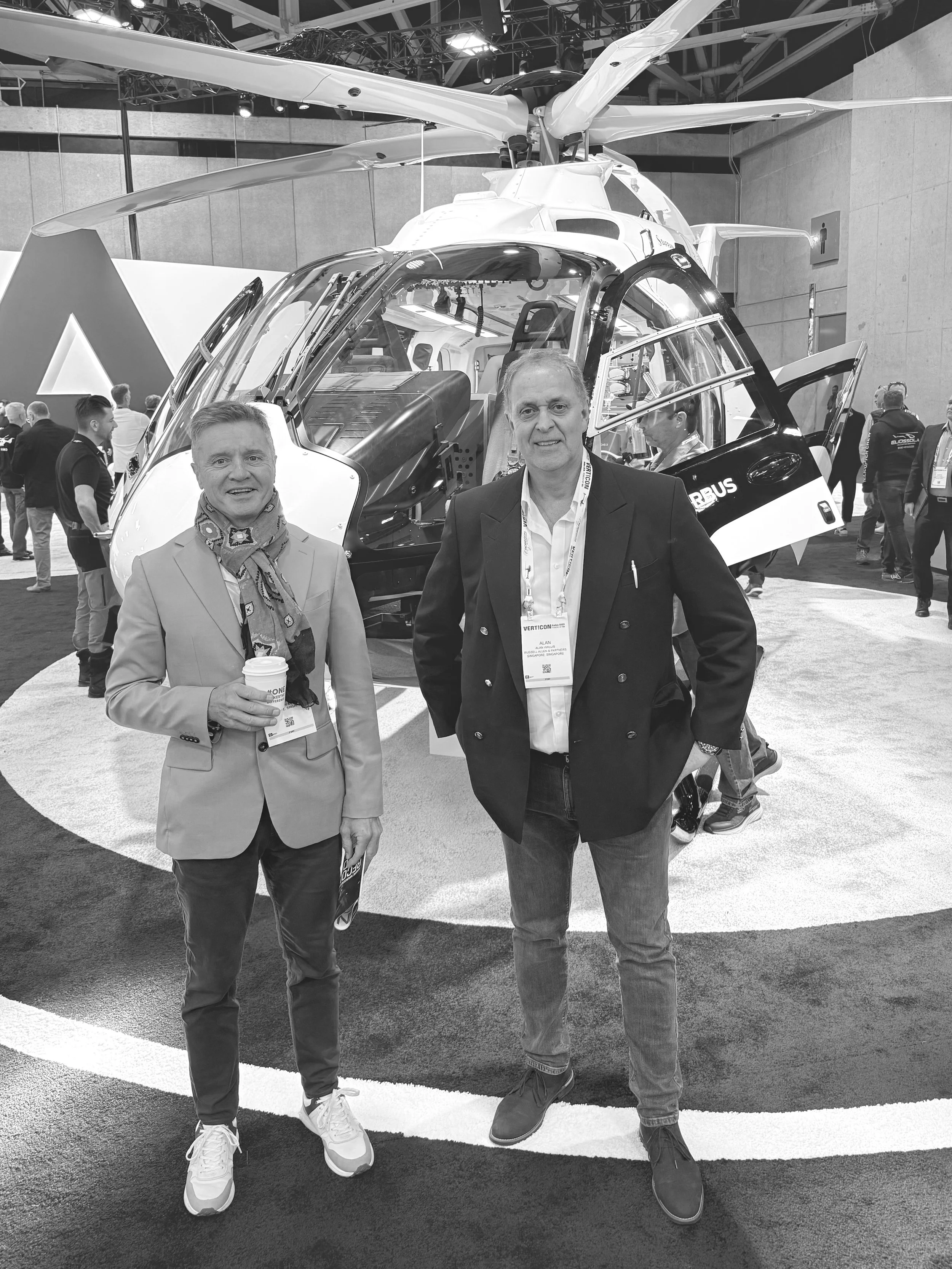 Two men standing in front of a helicopter at an exhibition. The man on the left is holding a coffee cup, and both are wearing conference badges.