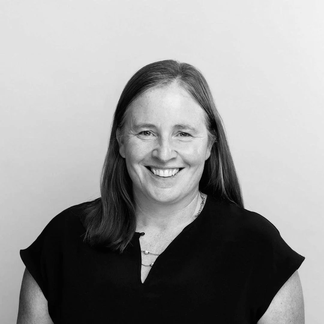 A smiling woman with shoulder-length hair wearing a black top posing in front of a plain background.