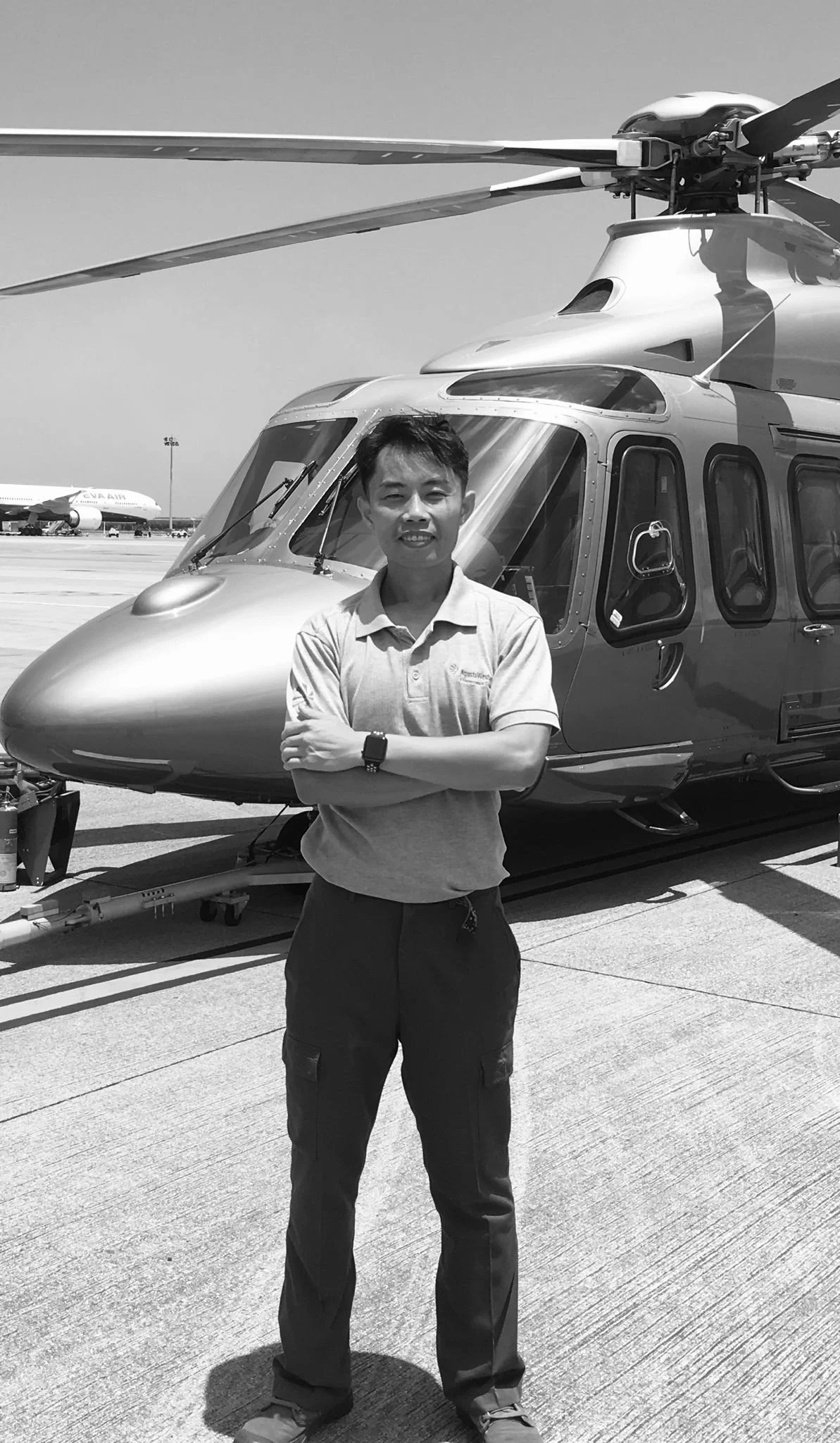 A person standing in front of a helicopter on an airport tarmac, with an airplane in the background.