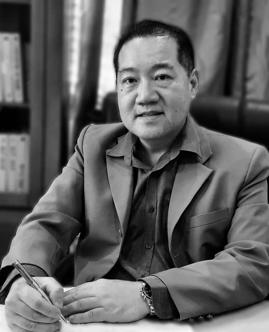 Black and white photo of a man in a suit sitting at a desk with papers, holding a pen, in an office setting.